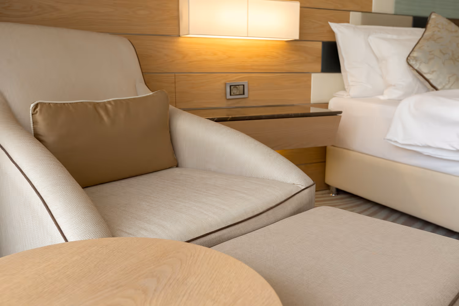 Bež fotelja sa svijetlosmeđim jastukom, mala stona lampa na zidu i krevet sa bijelim posteljinama u pozadini.