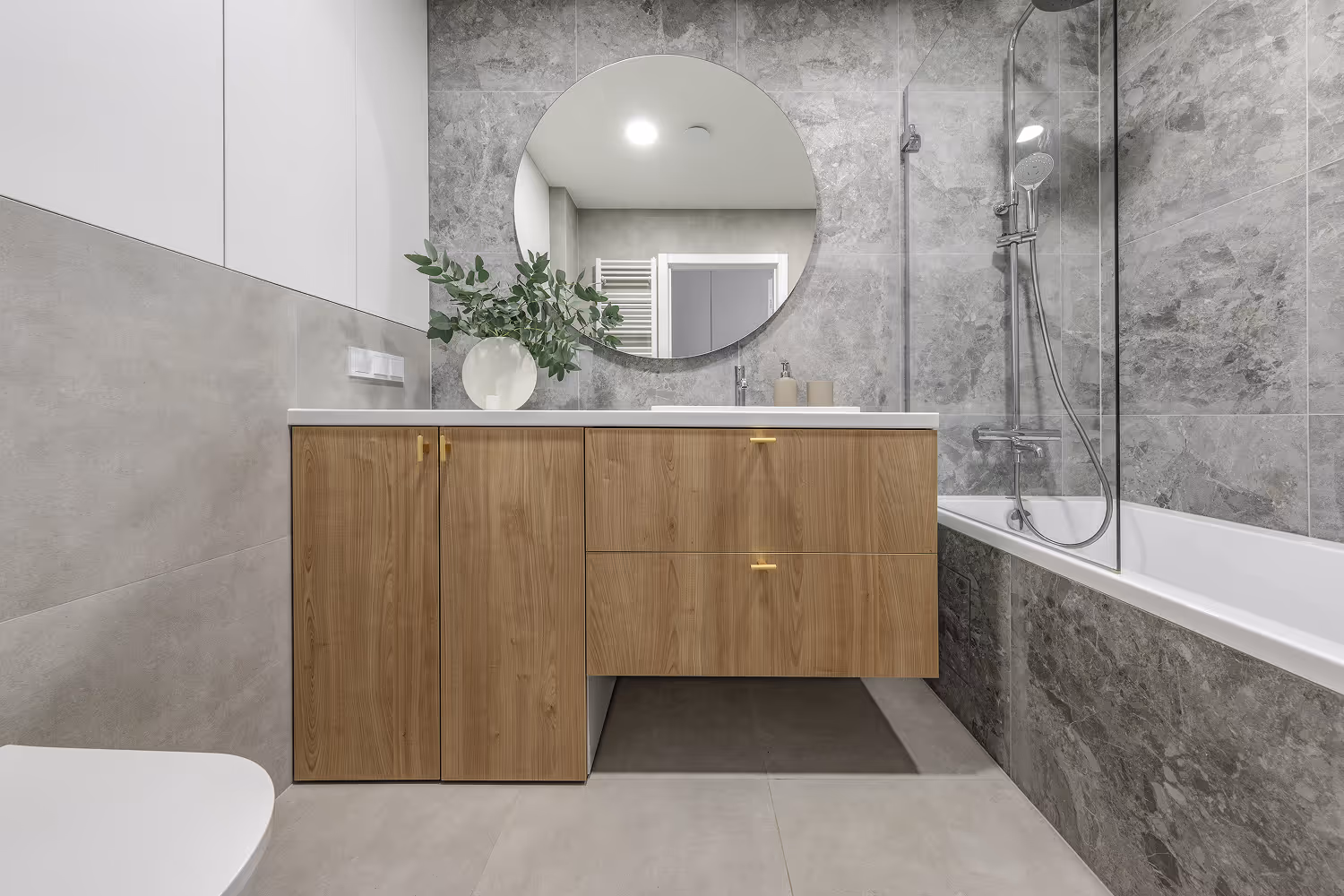 Modern bathroom with gray marble tiles, a wooden vanity with gold handles, round mirror, white sink, and a bathtub with a glass shower screen.