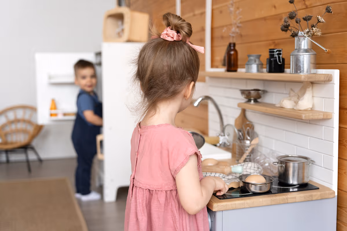 Djevojčica u ružičastoj haljini igra se za dječijom kuhinjom dok dječak stoji u pozadini sobe.