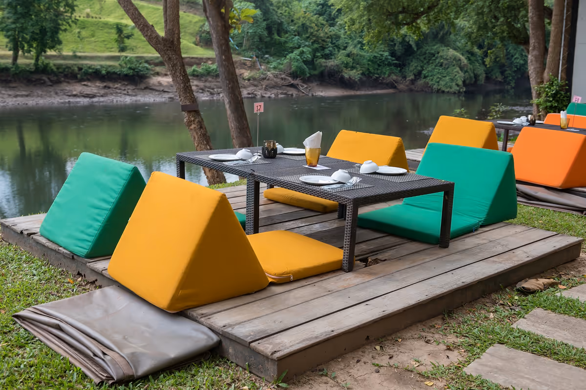 Na drvenoj platformi u vrtu uz rijeku postavljeni su stolovi s jastucima u zelenoj, žutoj i narandžastoj boji za sedenje blizu tla.