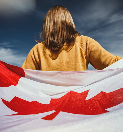 Better Flags | CiClO Flags | Person with brown hair and a mustard sweater holding a large Canadian flag against a cloudy sky.