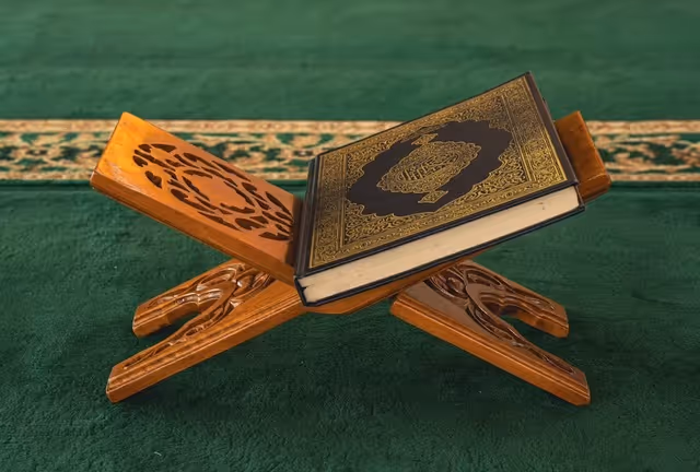 Un Coran avec une reliure décorative dorée posé sur un lutrin en bois sculpté, sur une moquette verte.