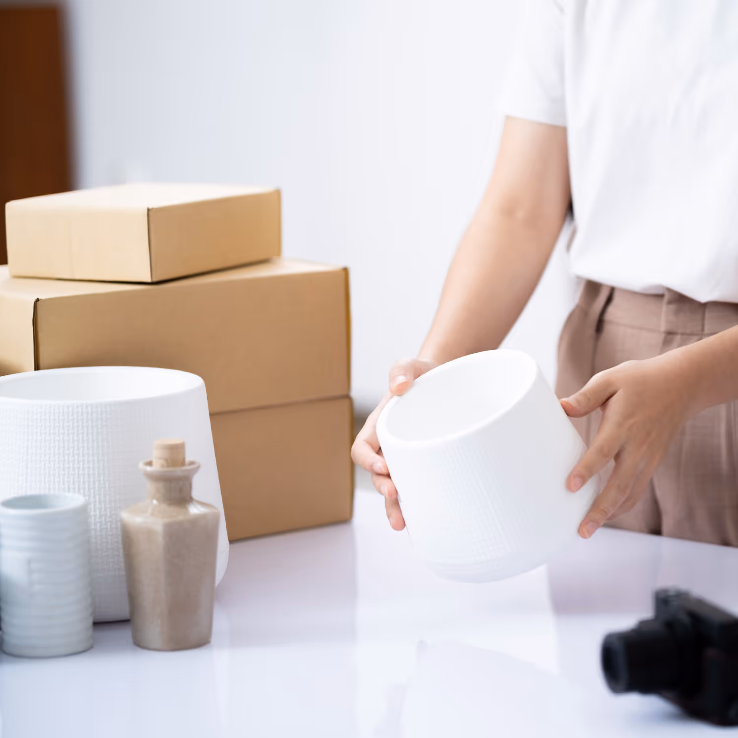 Person holding a white textured cylindrical pot with cardboard boxes and ceramic bottles on a table.