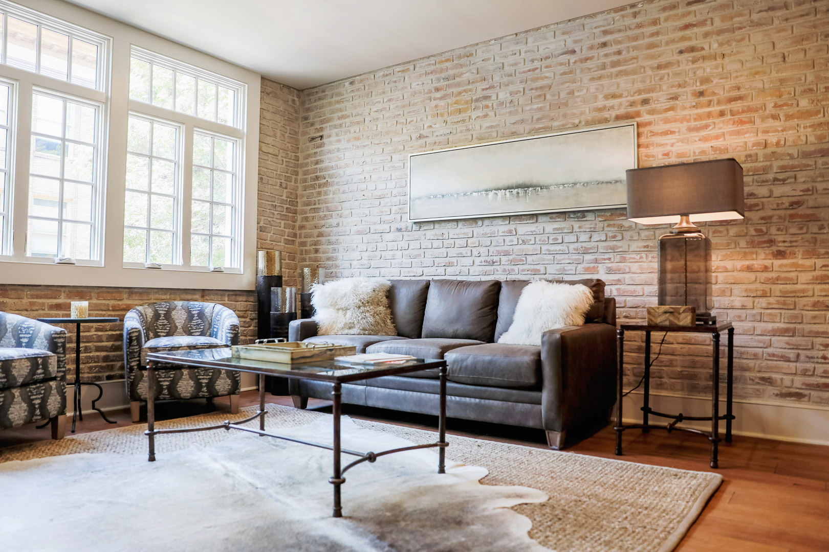 Modern living room with a dark leather sofa topped with white fluffy pillows, patterned armchairs, a glass coffee table, and a side table with a lamp against a whitewashed brick wall.