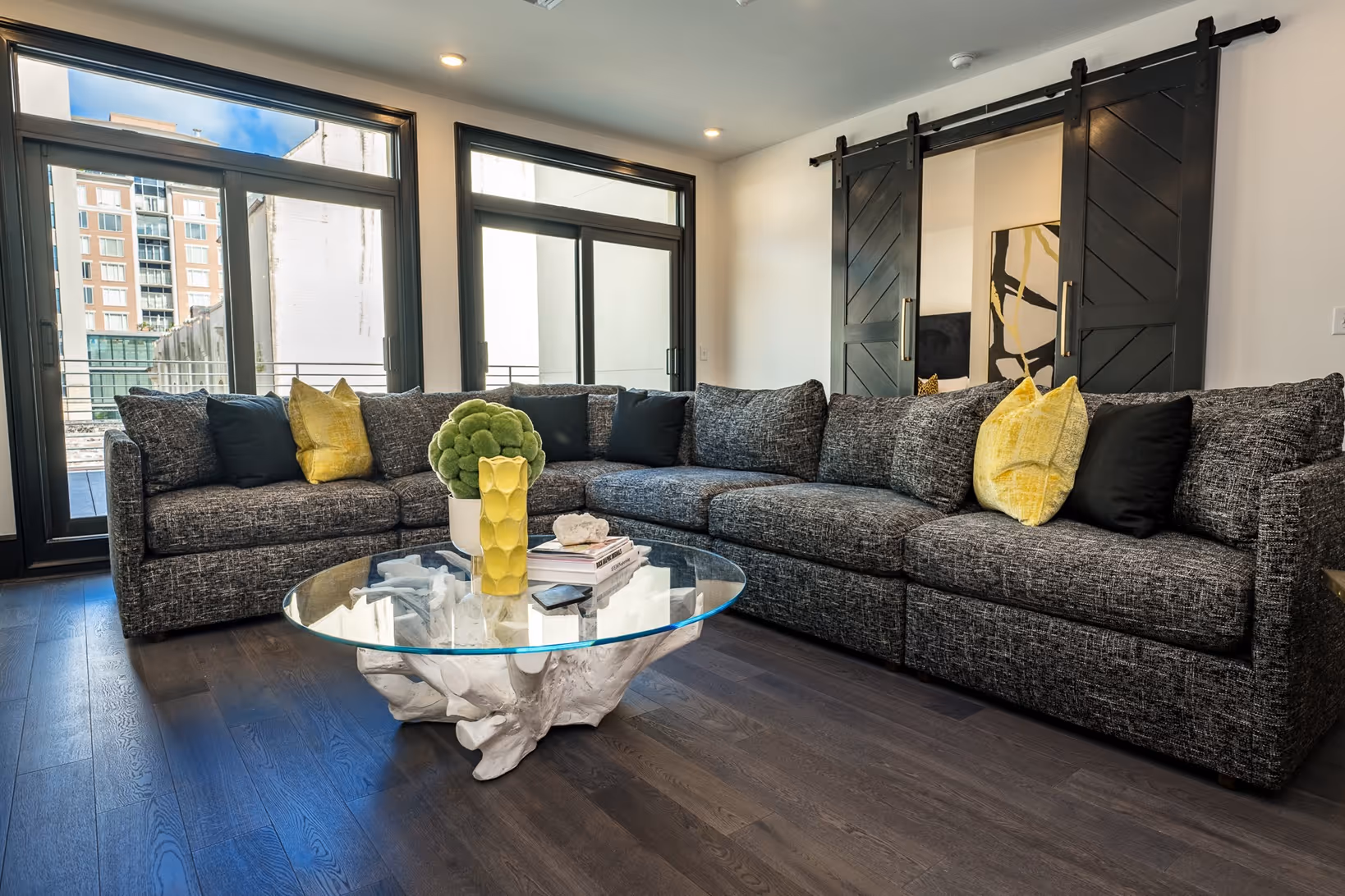 Modern living room with gray sectional sofa, yellow and black pillows, glass coffee table with white sculptural base, and large windows with city view.
