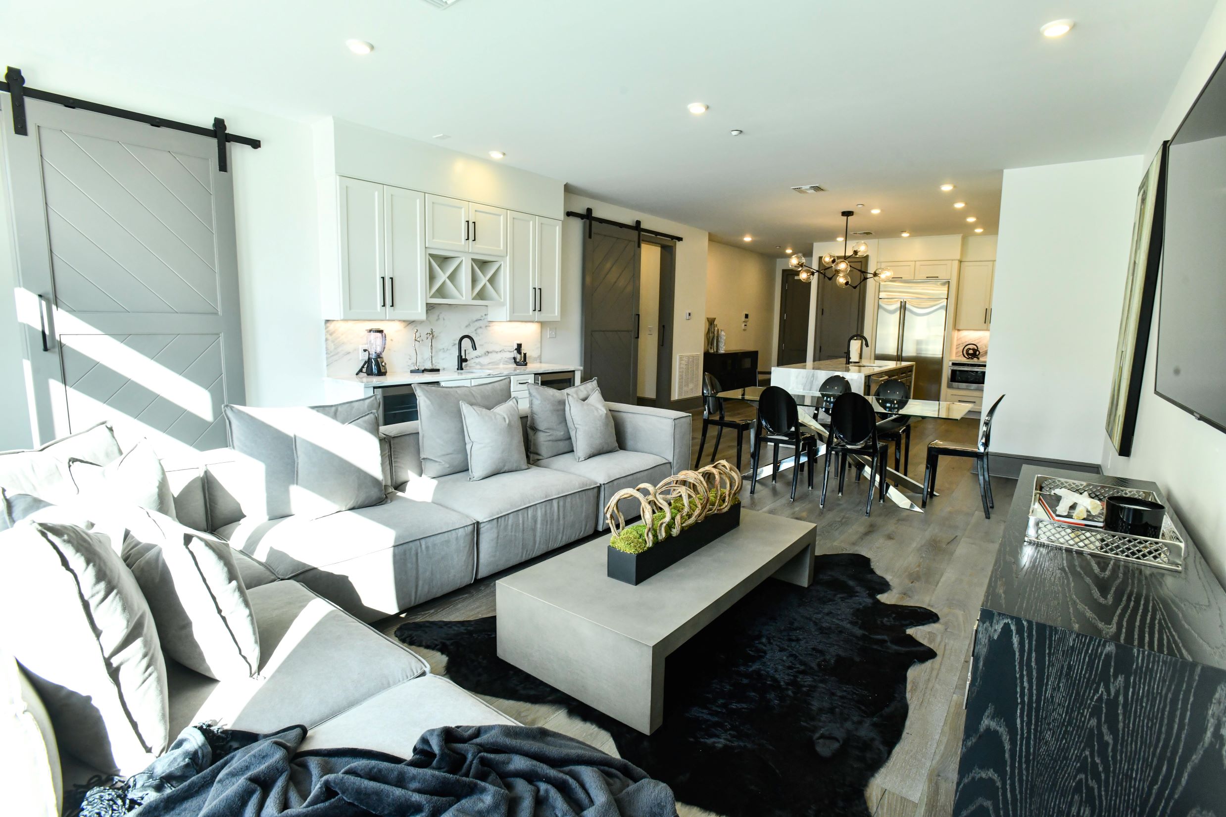 Modern open living room with gray sectional sofa, concrete coffee table, black rug, glass dining table with black chairs, and white kitchen with stainless steel appliances.