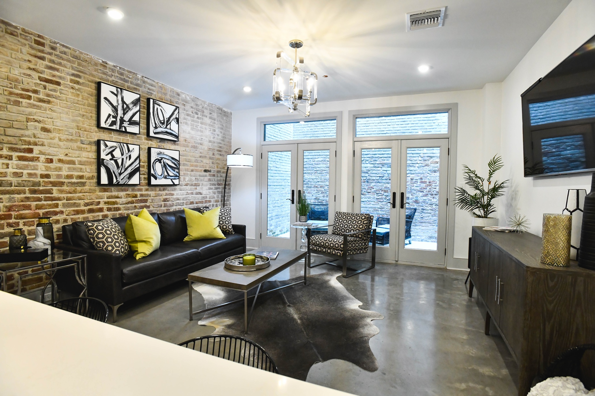 Modern living room with exposed brick wall, black leather sofa with yellow and patterned cushions, patterned armchair, glass coffee table on cowhide rug, and large glass doors.
