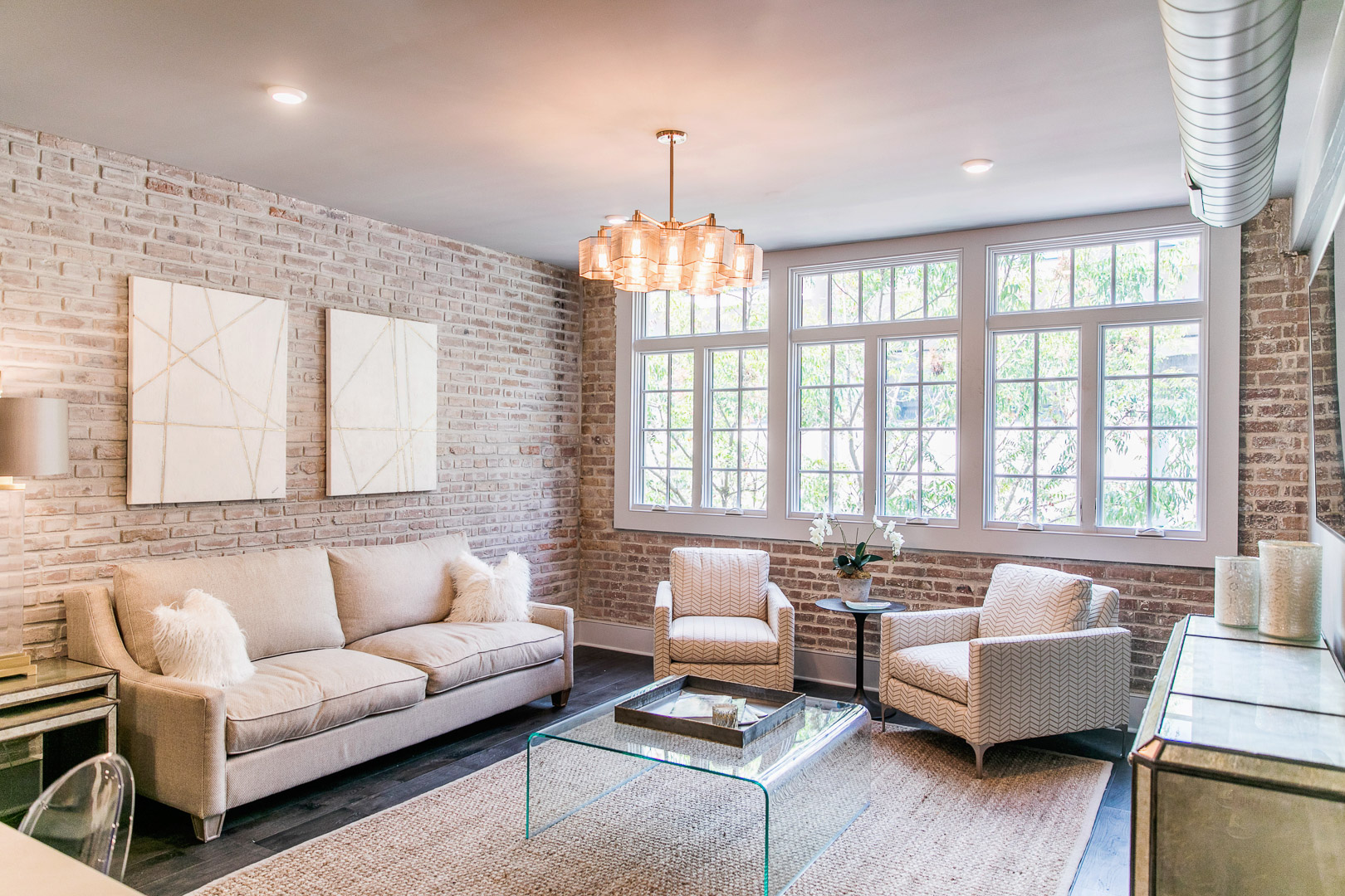 Bright living room with beige sofa, two patterned armchairs, glass coffee table, and large windows on exposed brick walls.