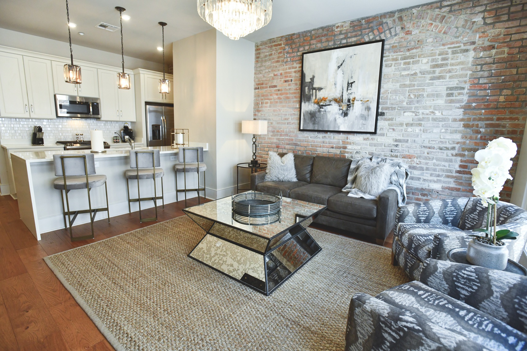 Modern living room with exposed brick wall, gray sofa with fluffy pillows, mirrored coffee table, patterned chairs, and kitchen with bar stools and pendant lights.