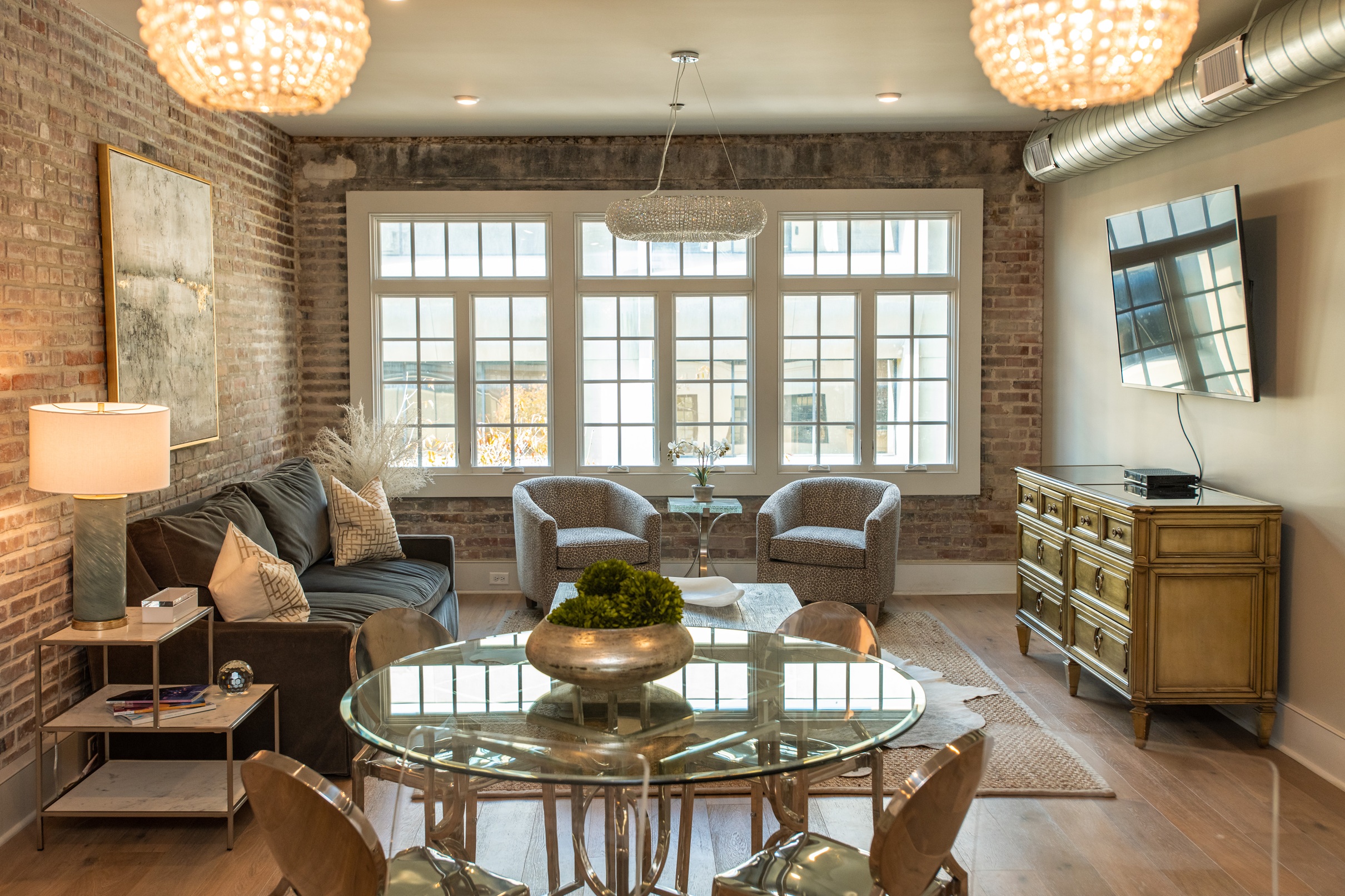 Modern living room with exposed brick walls, a gray sofa with pillows, two patterned armchairs, a glass dining table with chairs, a wooden sideboard, and large windows.