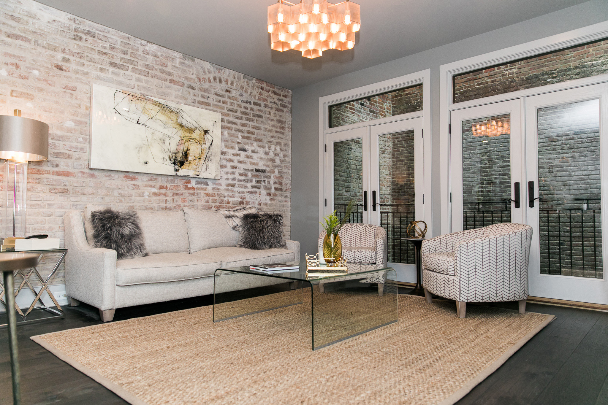 Modern living room with a beige sofa, two patterned armchairs, glass coffee table on a woven rug, exposed brick wall, and large glass doors.