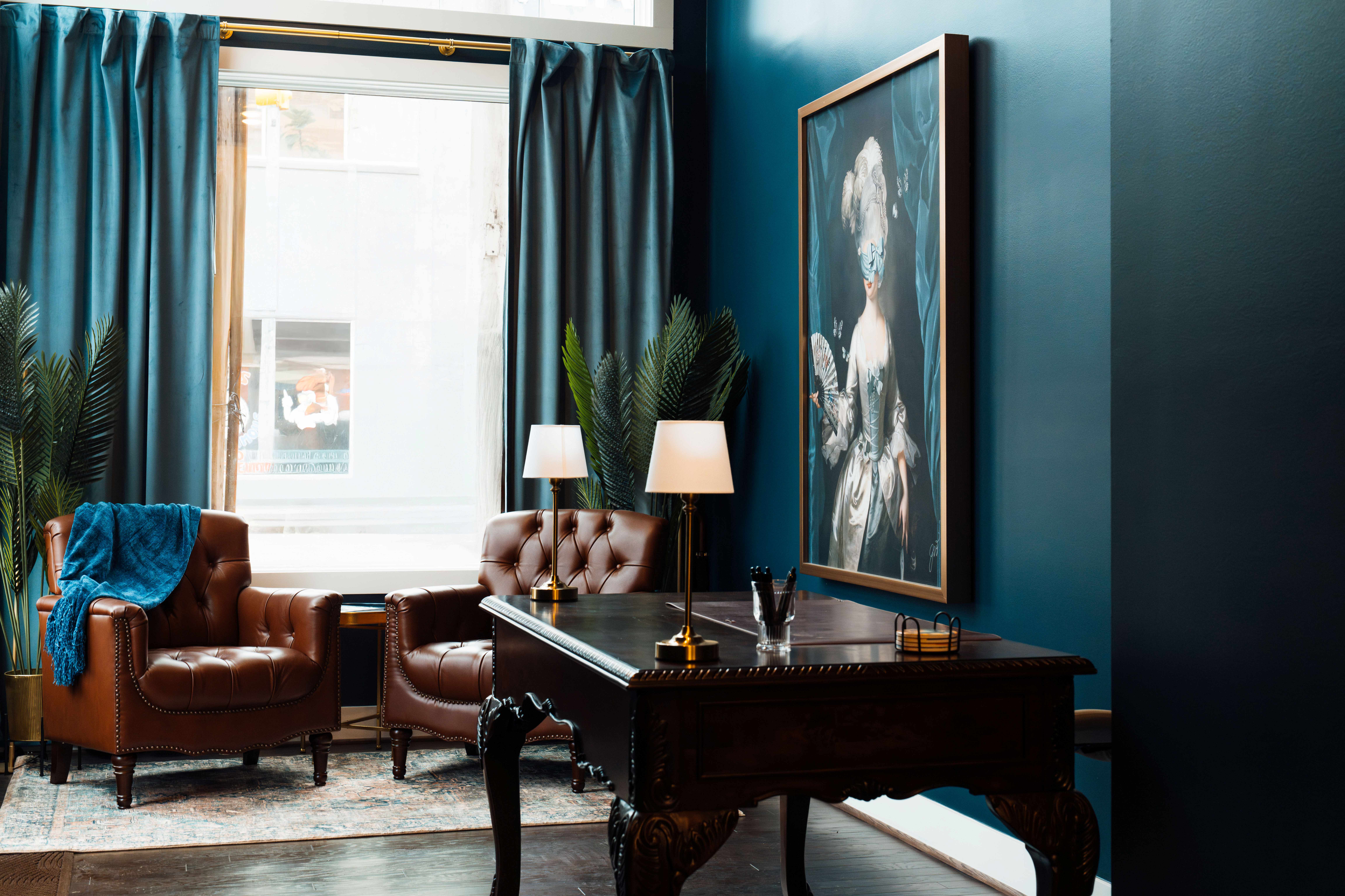 Cozy sitting area with two brown leather armchairs, blue curtains, a dark wooden desk with lamps, and a framed portrait on teal wall.
