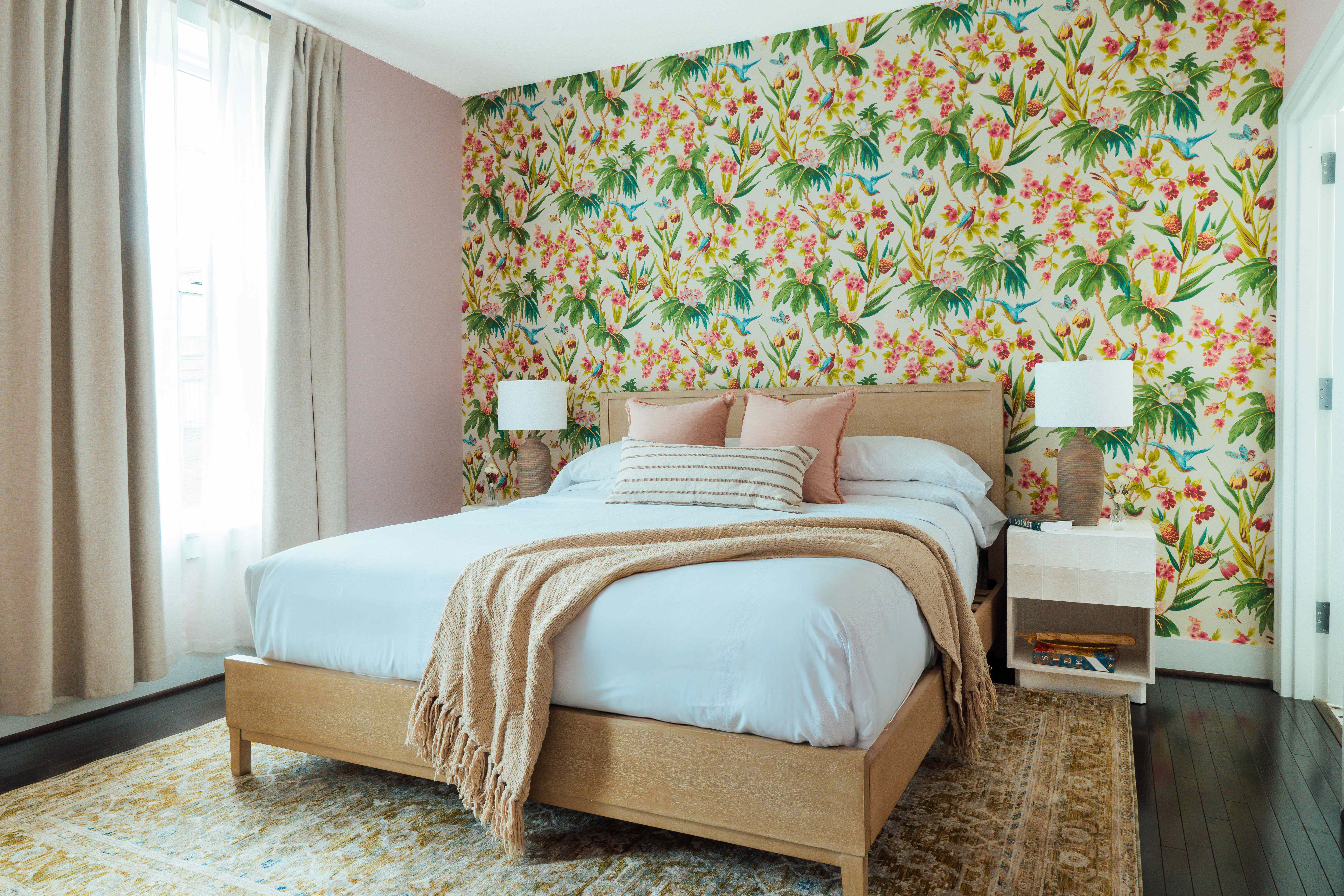 Bedroom with wooden bed featuring white bedding, beige throw, pink and striped pillows, floral wallpaper, two bedside tables with lamps, and tall beige curtains.