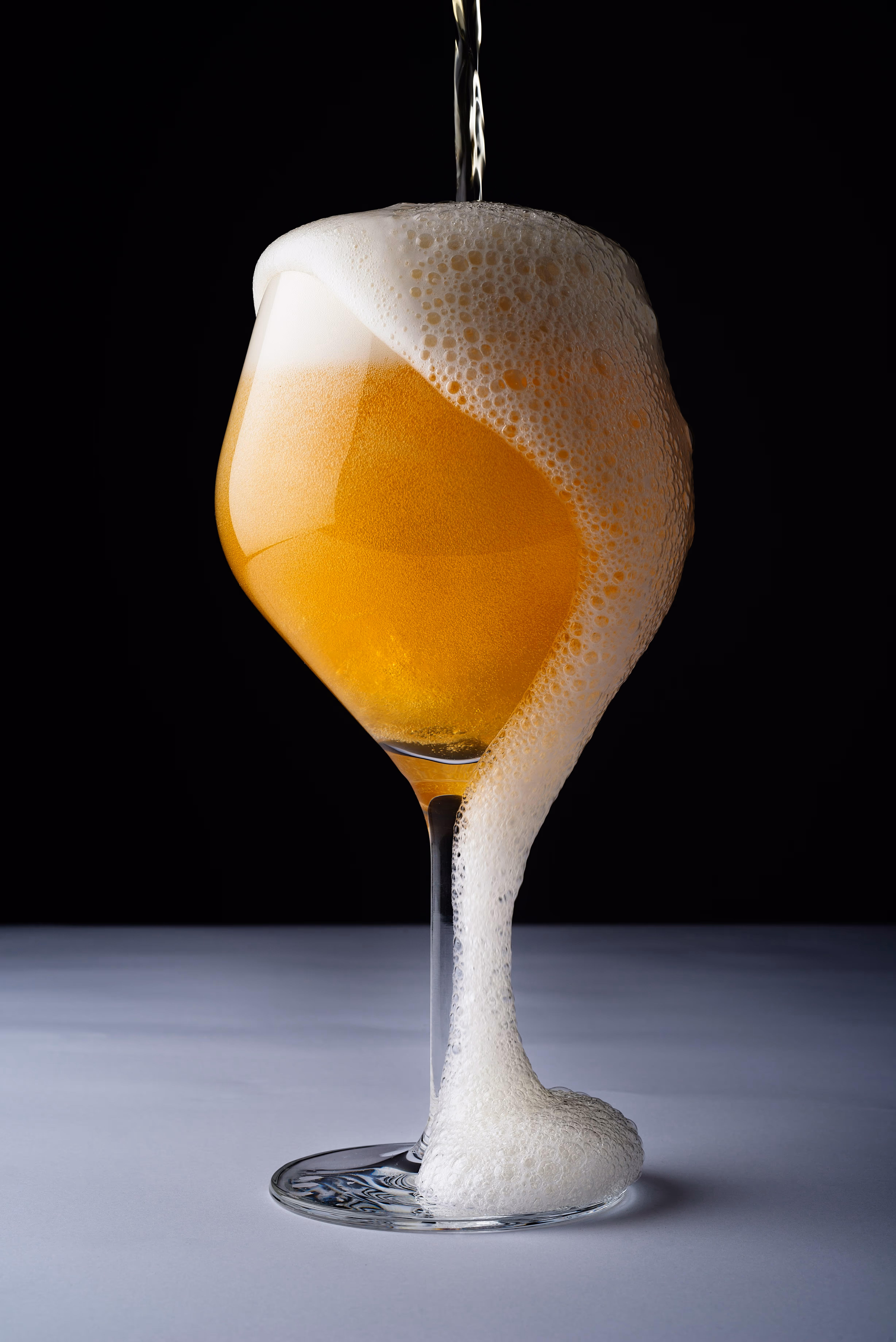 Glass of beer being poured with foam overflowing down the side onto a white surface with black background.