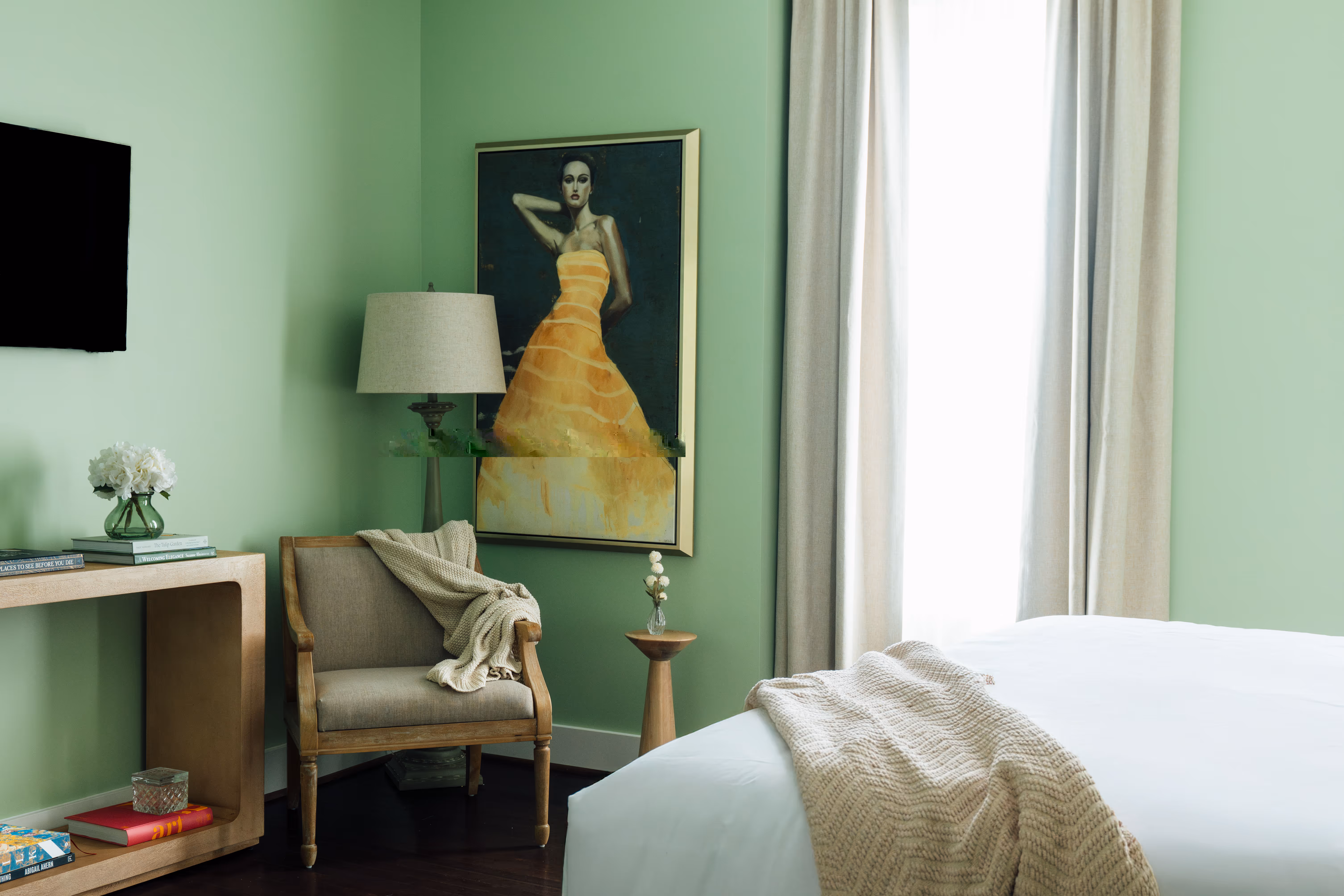 Cozy bedroom corner with beige armchair draped with a knit blanket, a floor lamp, a framed painting of a woman in a yellow dress, and a wooden side table with a small vase.