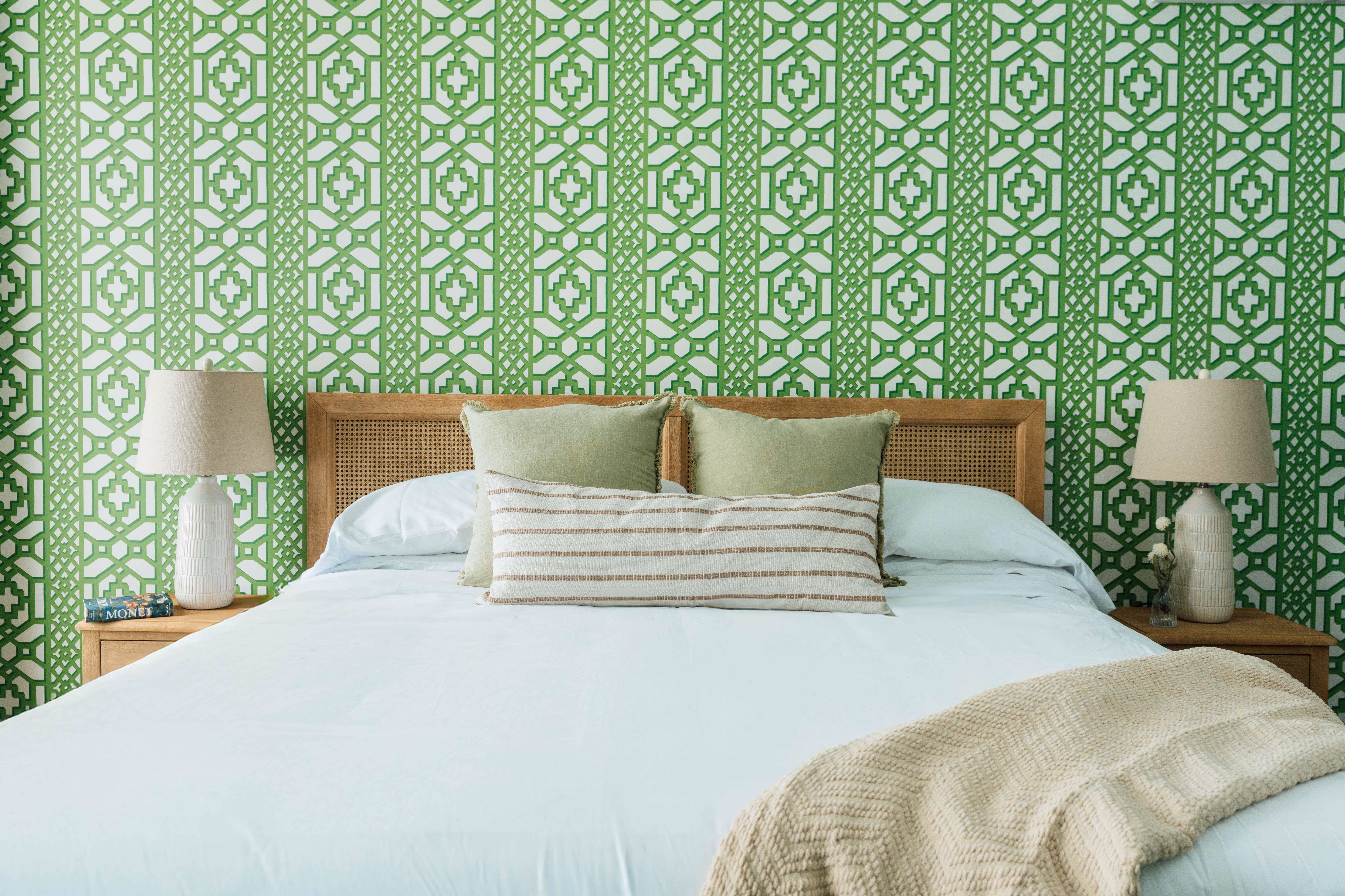 Bed with white bedding, two green pillows, striped lumbar pillow, beige knitted throw, wooden headboard, and matching bedside tables with beige lamps in front of green patterned wallpaper.
