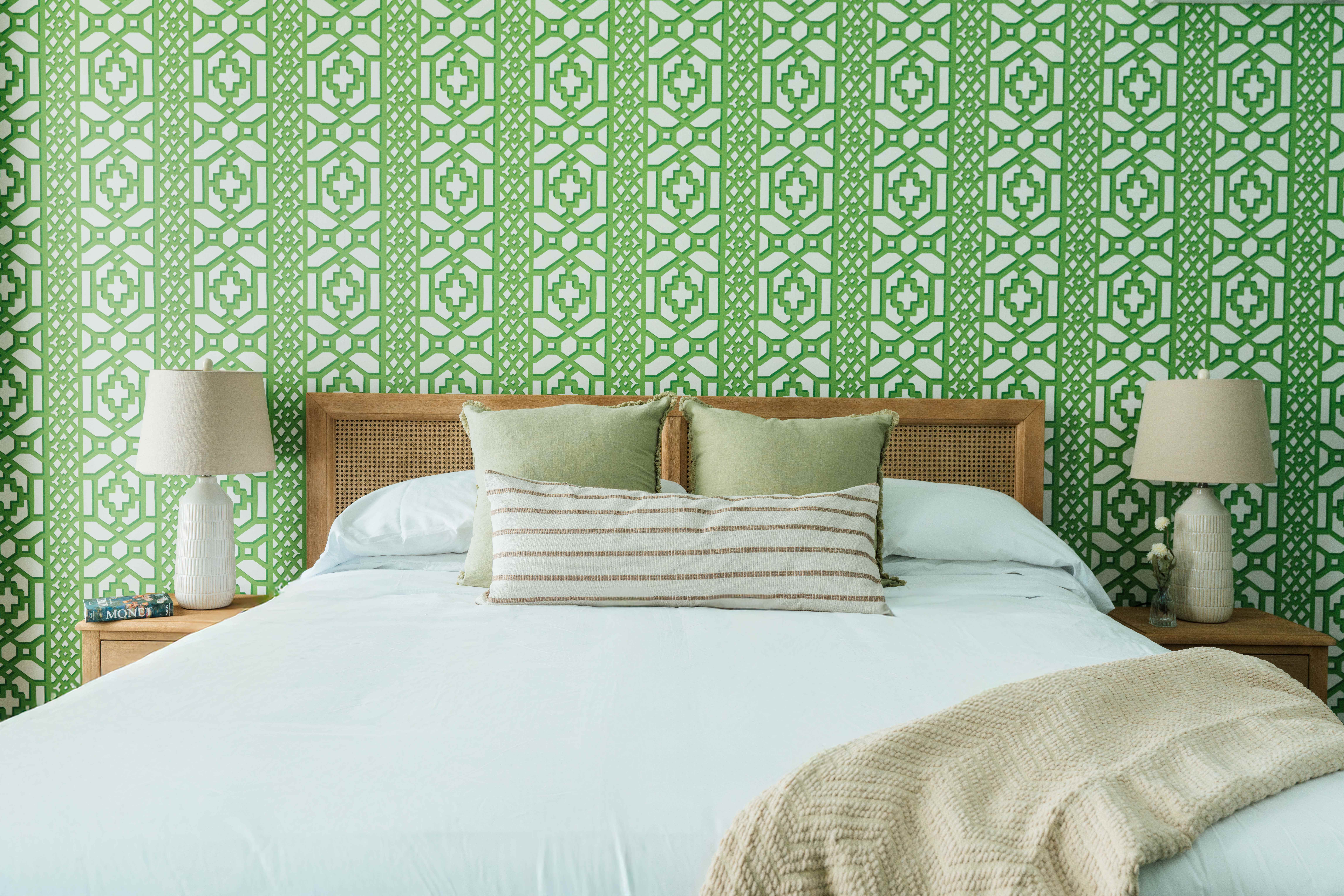 Bed with white bedding, two green pillows, striped lumbar pillow, beige knitted throw, wooden headboard, and matching bedside tables with beige lamps in front of green patterned wallpaper.