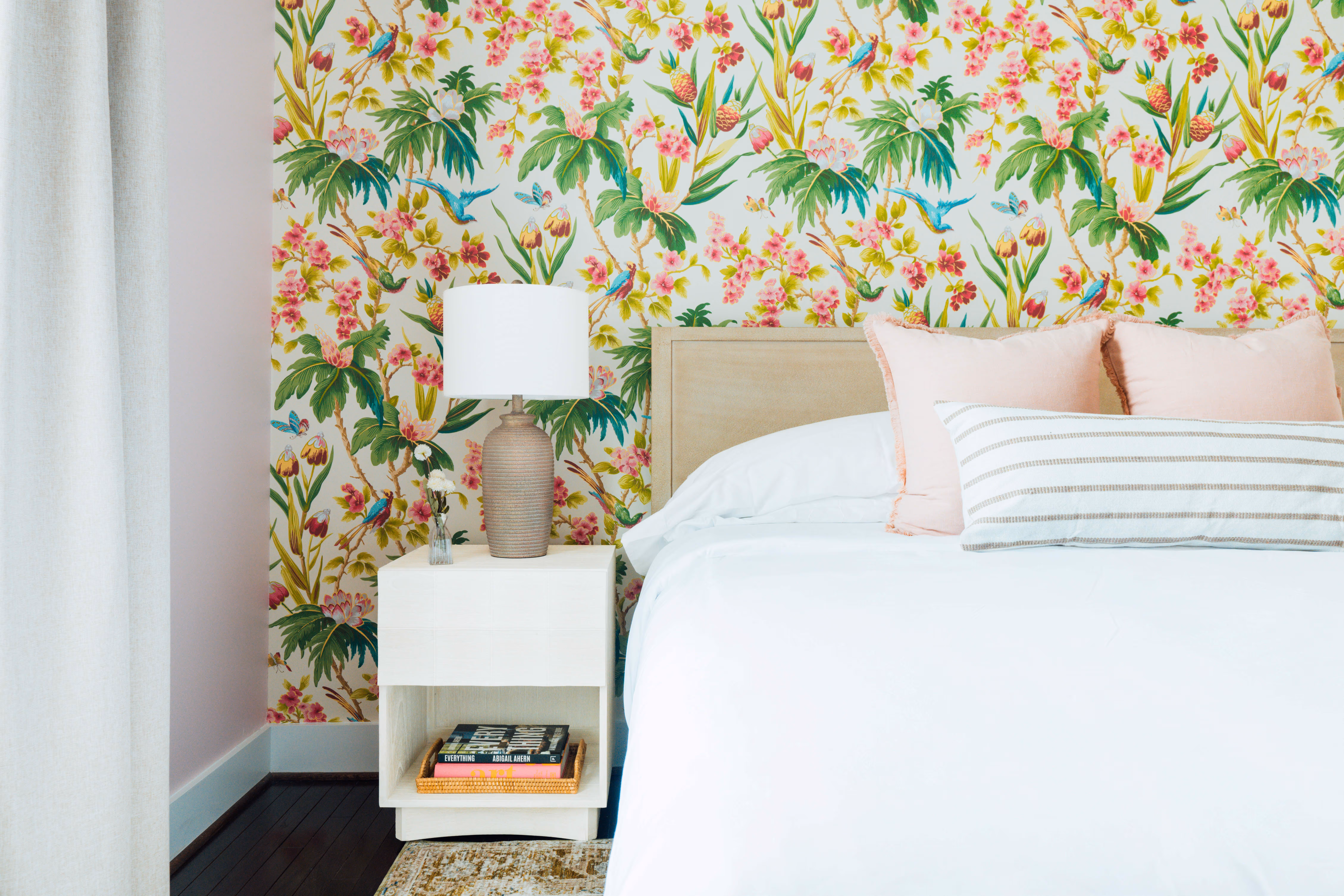 Bedroom corner with a white bed, light pink and striped pillows, floral wallpaper, and a nightstand holding a lamp and a small vase.