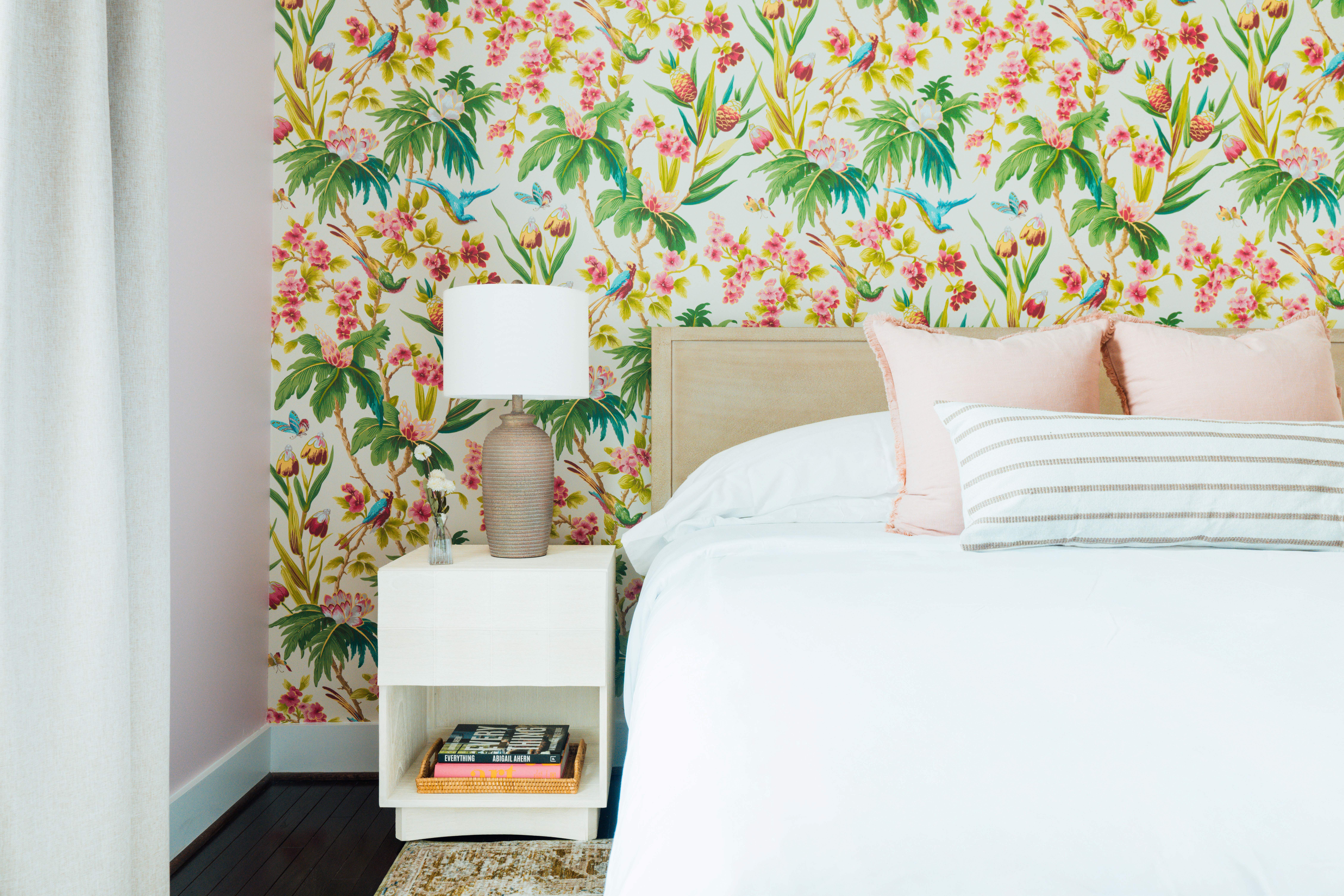 Bedroom corner with a white bed, light pink and striped pillows, floral wallpaper, and a nightstand holding a lamp and a small vase.