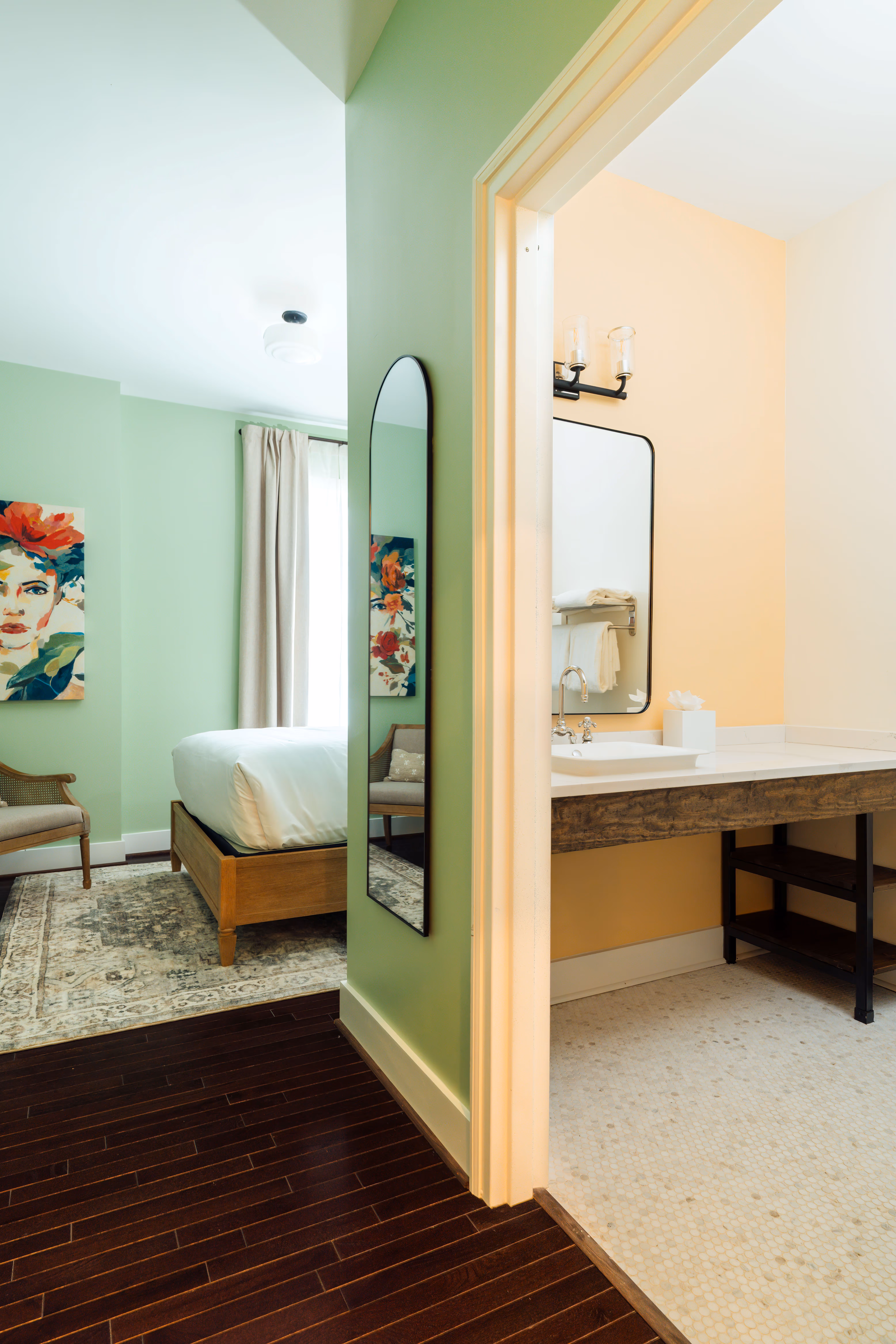 View from dark wood floor hallway into a bedroom with green walls and a bathroom with light peach walls and white tiled floor.