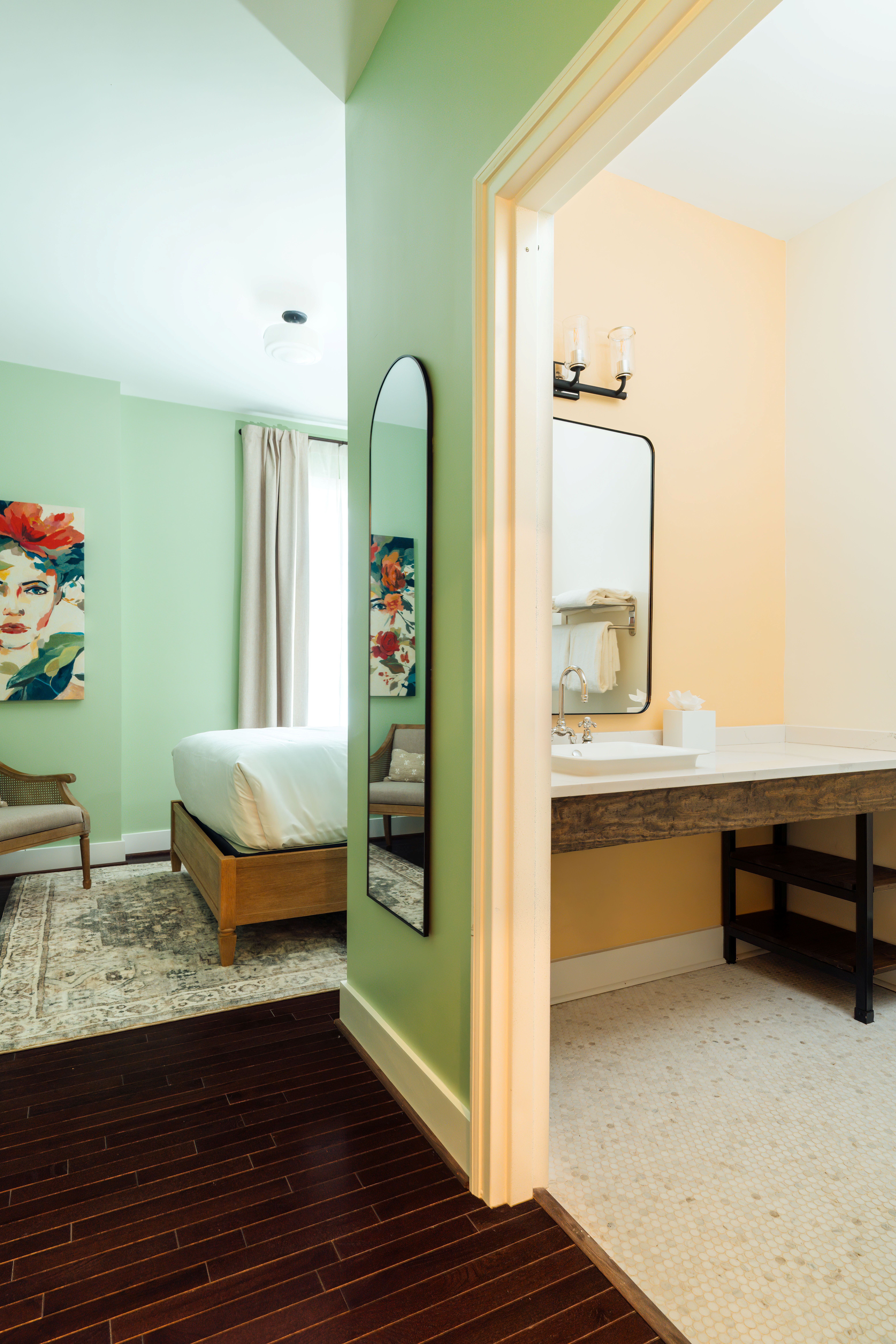View from dark wood floor hallway into a bedroom with green walls and a bathroom with light peach walls and white tiled floor.