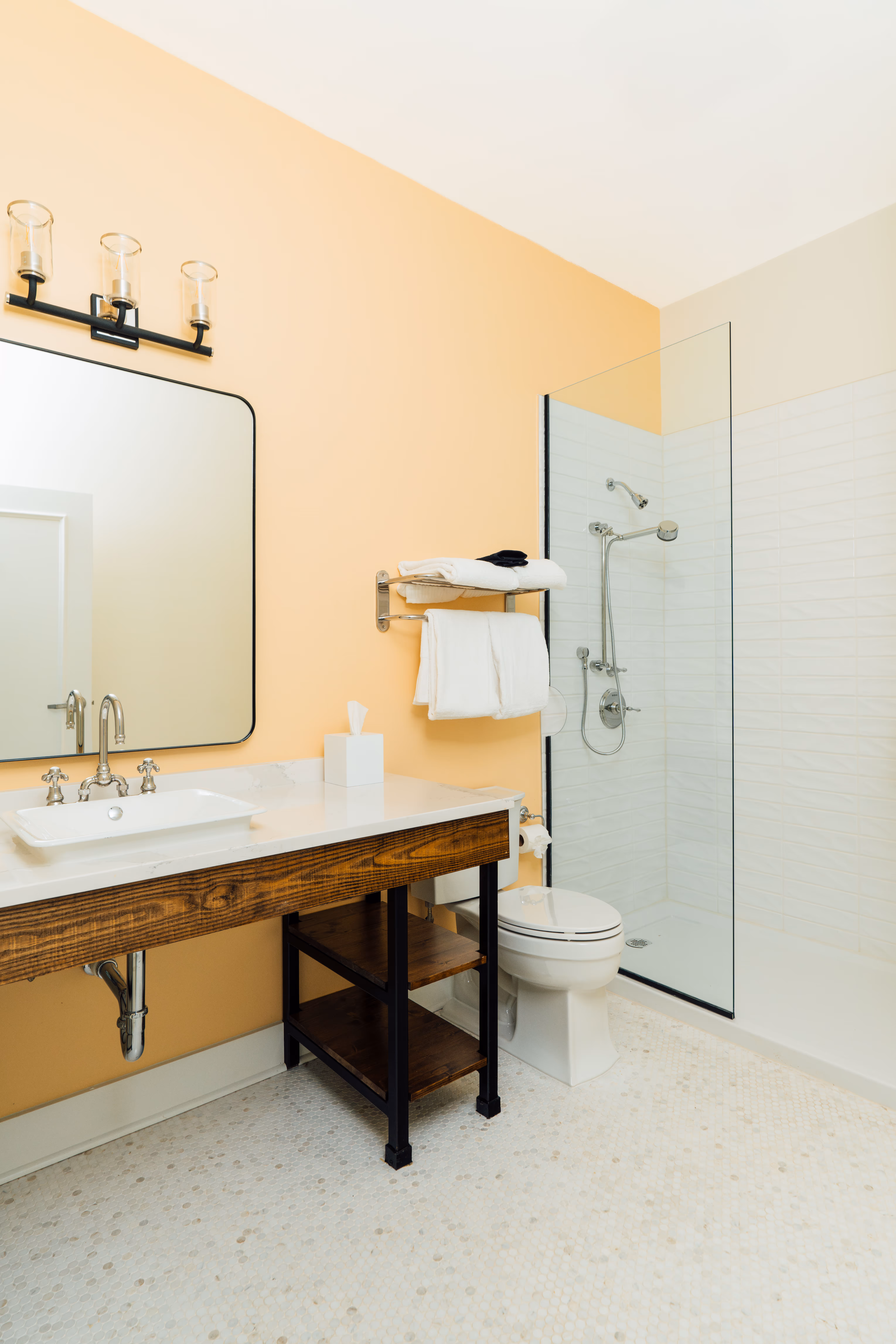 Modern bathroom with tan walls, a wooden vanity with white sink, a large mirror, a glass shower enclosure, and a toilet with folded white towels on a wall rack.