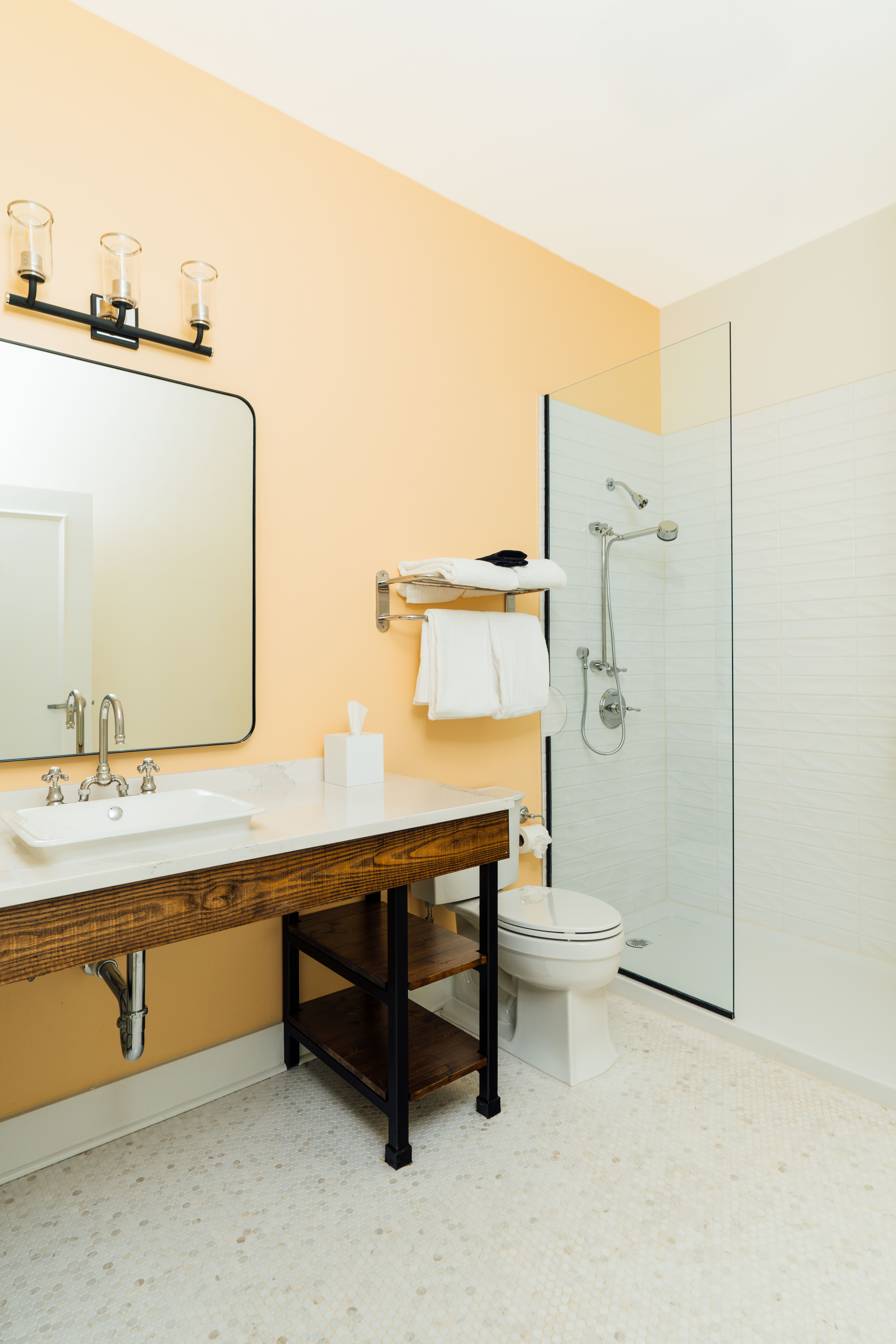 Modern bathroom with tan walls, a wooden vanity with white sink, a large mirror, a glass shower enclosure, and a toilet with folded white towels on a wall rack.