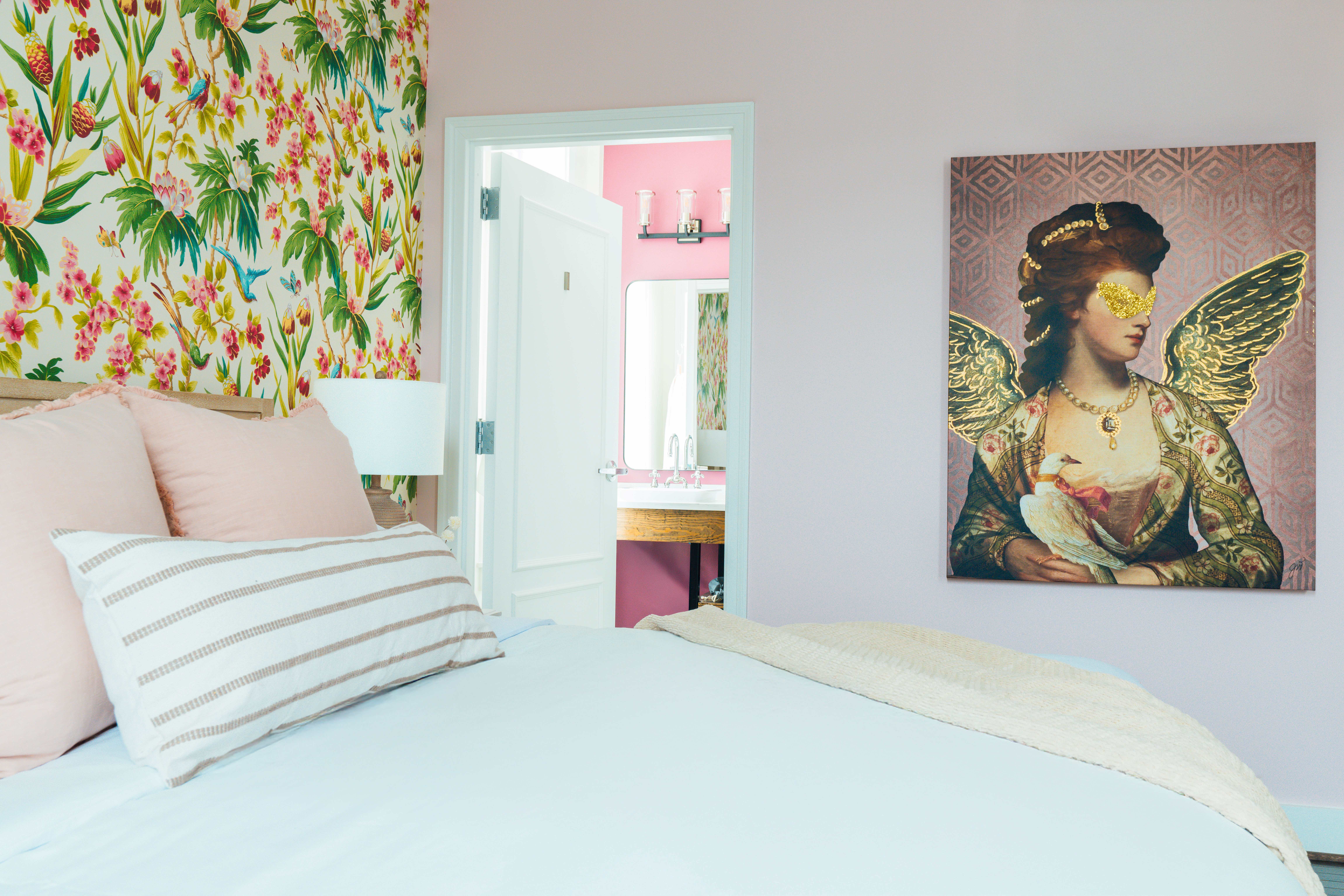 Bedroom with floral wallpaper, a bed with pink and striped pillows, and a painting of a woman with golden wings and a bird on her arm on the wall.
