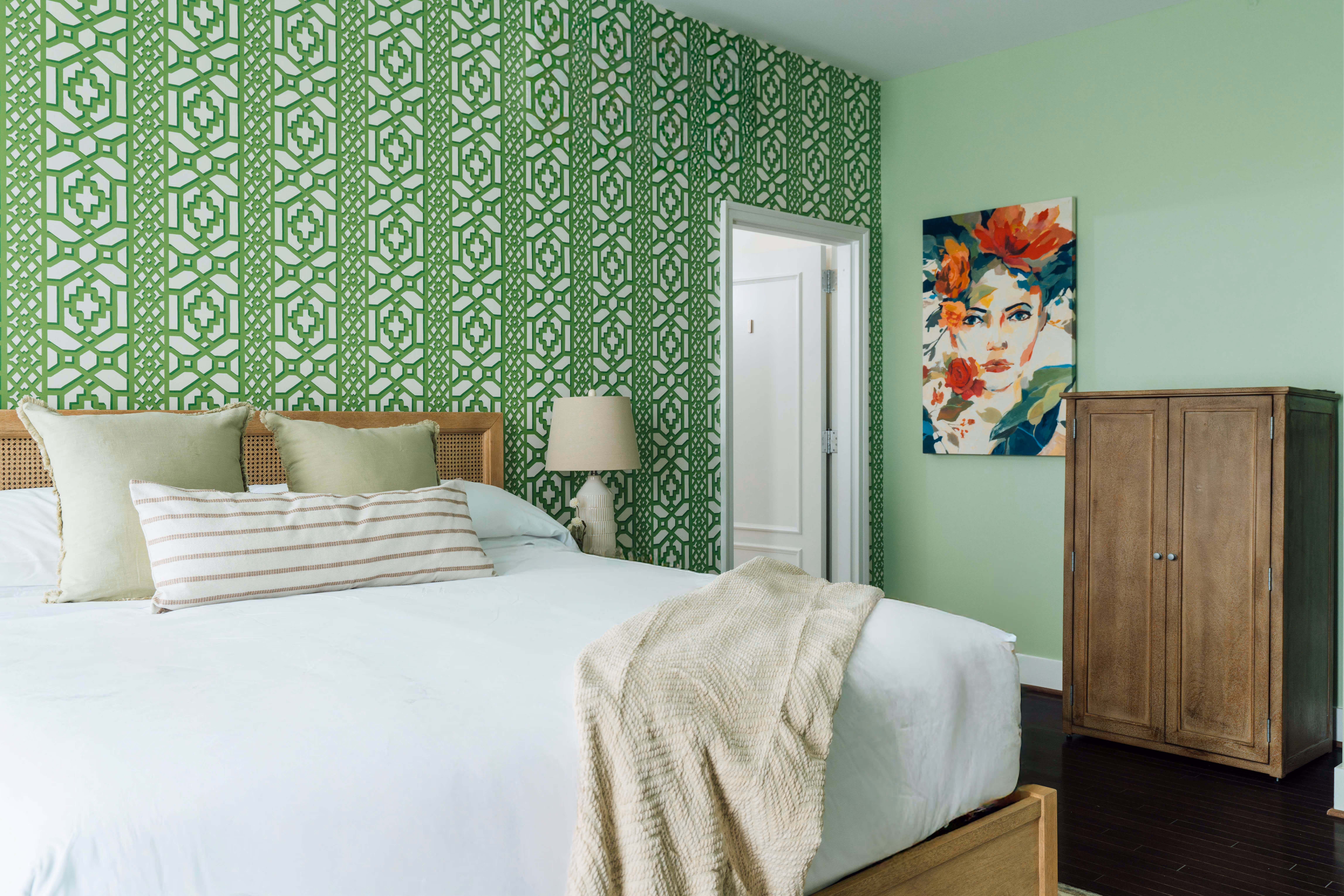 Bedroom with green geometric patterned accent wall, bed with light green pillows and cream blanket, wooden nightstand with lamp, floral portrait, and wooden wardrobe.