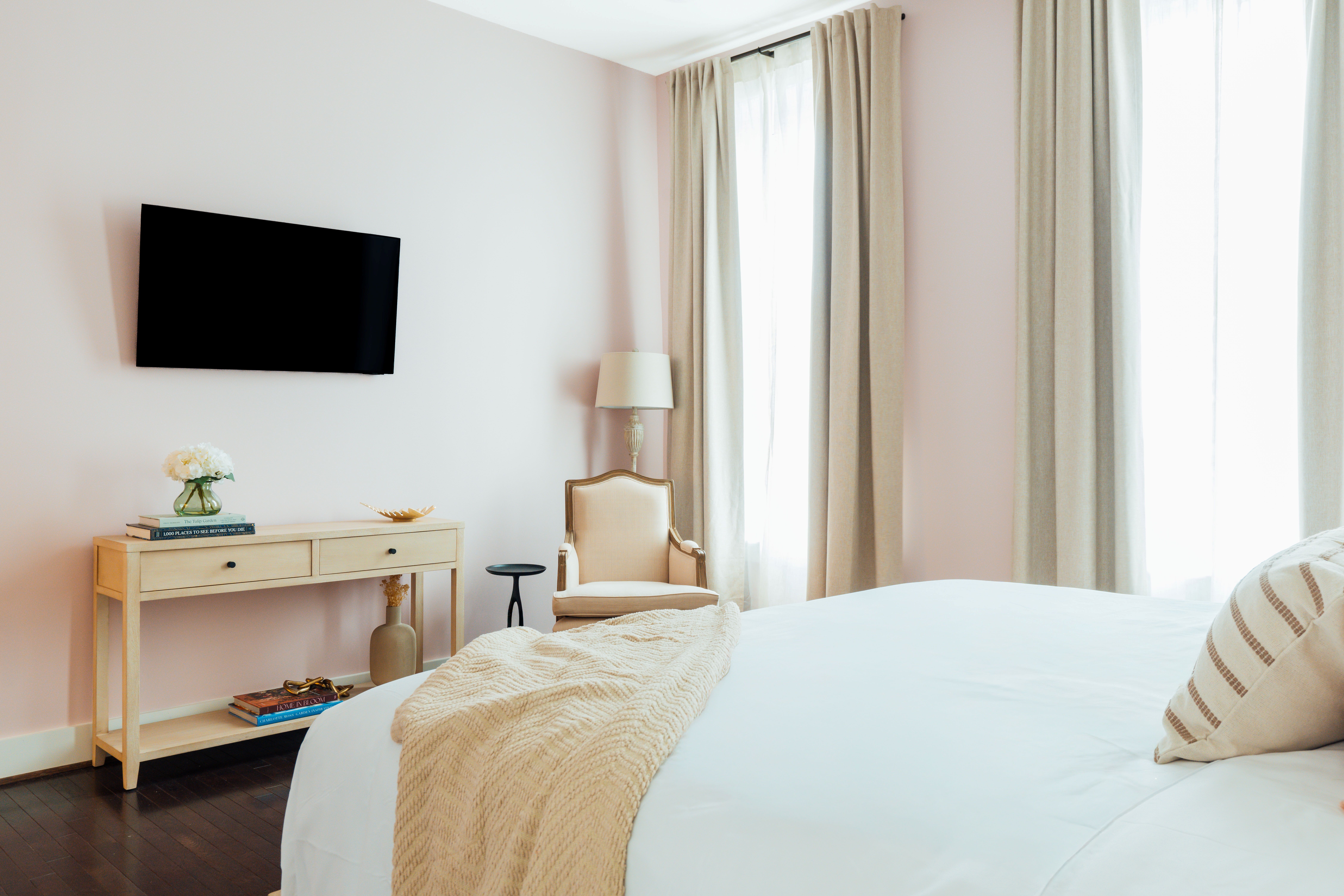 Bright bedroom corner with a beige armchair, wooden console table, wall-mounted TV, and bed with cream throw and striped pillow.