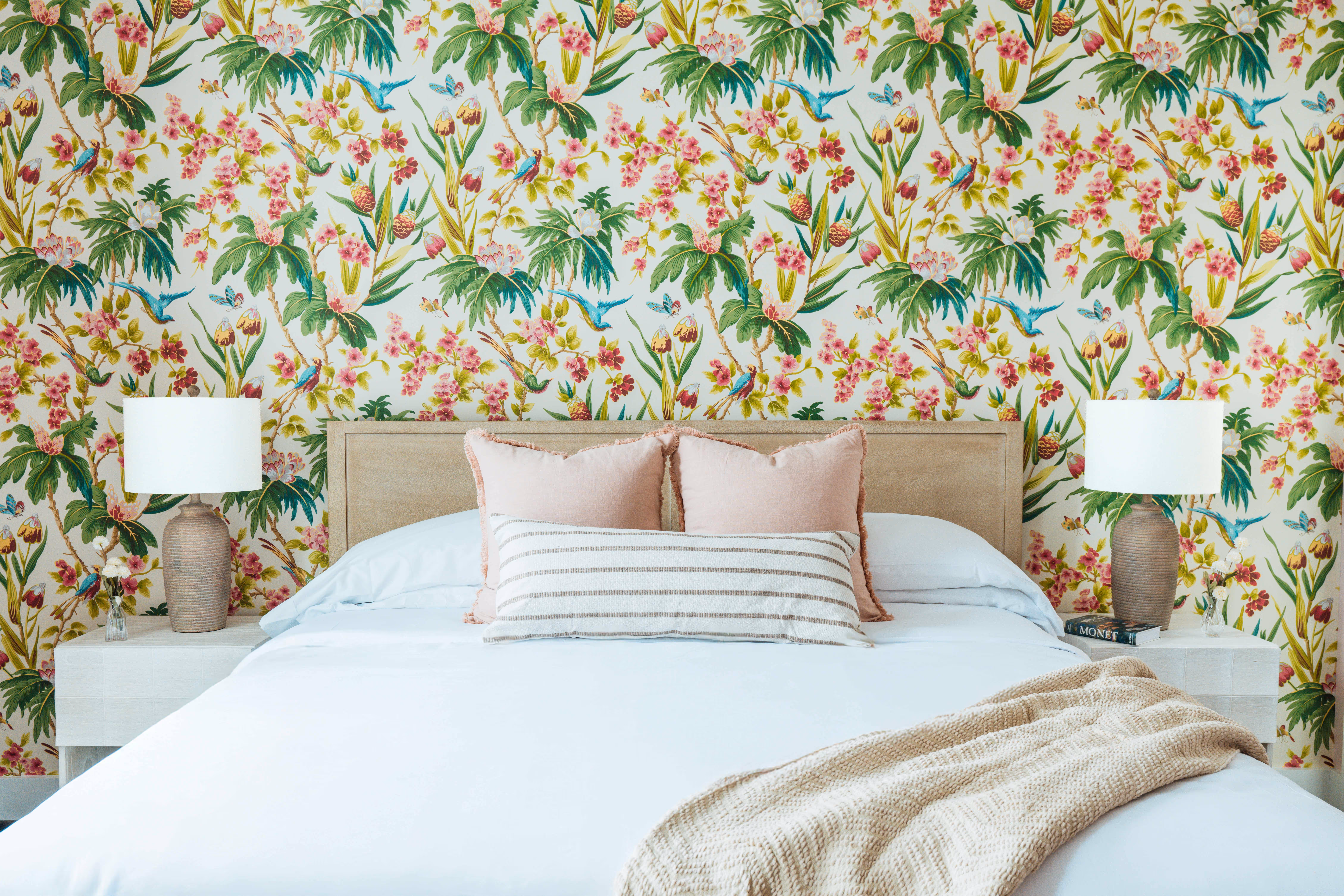 Bedroom with a beige headboard bed, white bedding, striped and pink pillows, beige knitted throw, floral wallpaper with birds, and matching bedside lamps on white nightstands.