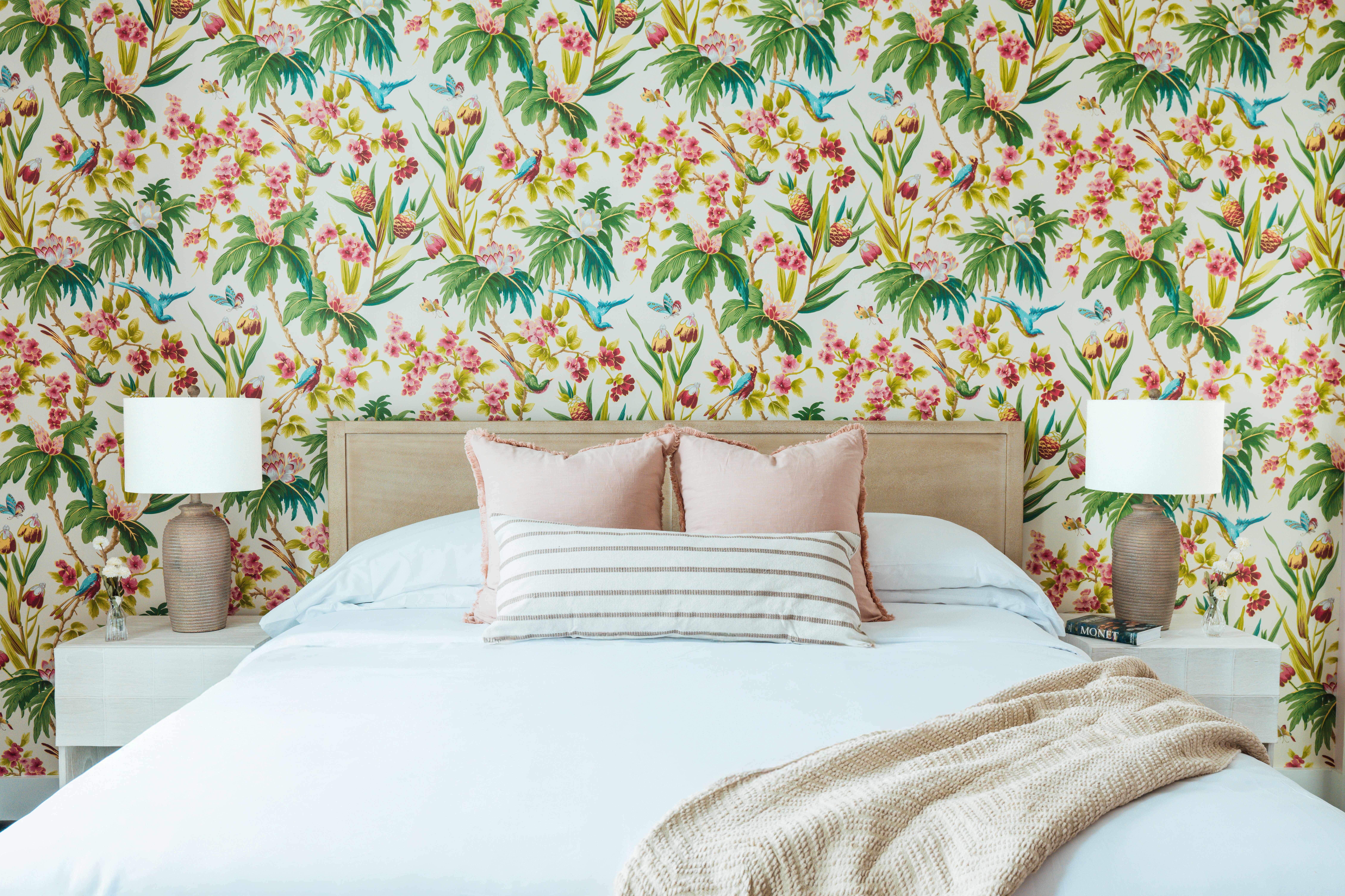 Bedroom with a beige headboard bed, white bedding, striped and pink pillows, beige knitted throw, floral wallpaper with birds, and matching bedside lamps on white nightstands.