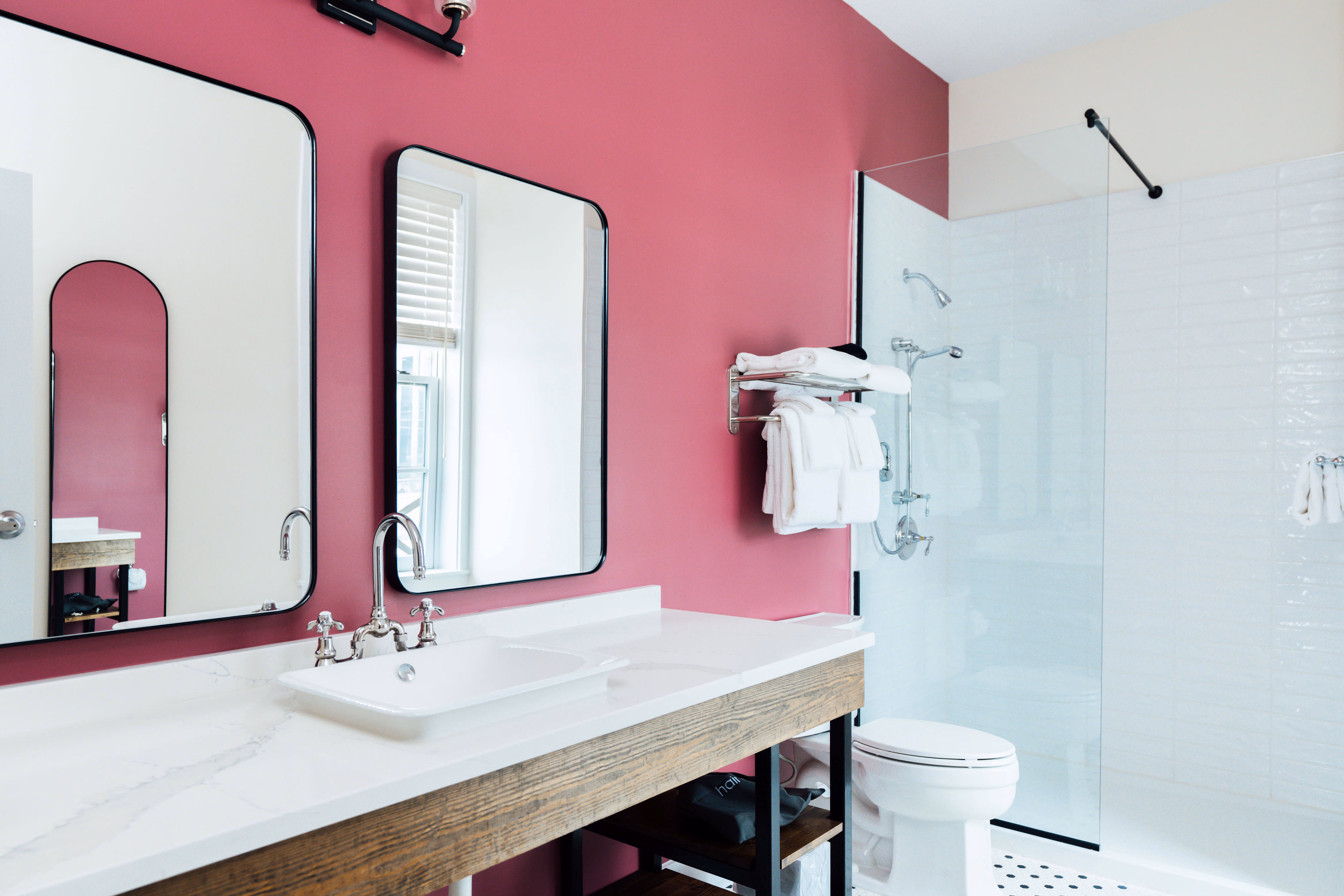 Bathroom with a pink accent wall, white sink on a wooden vanity, two mirrors, towel rack with white towels, and a glass shower enclosure.