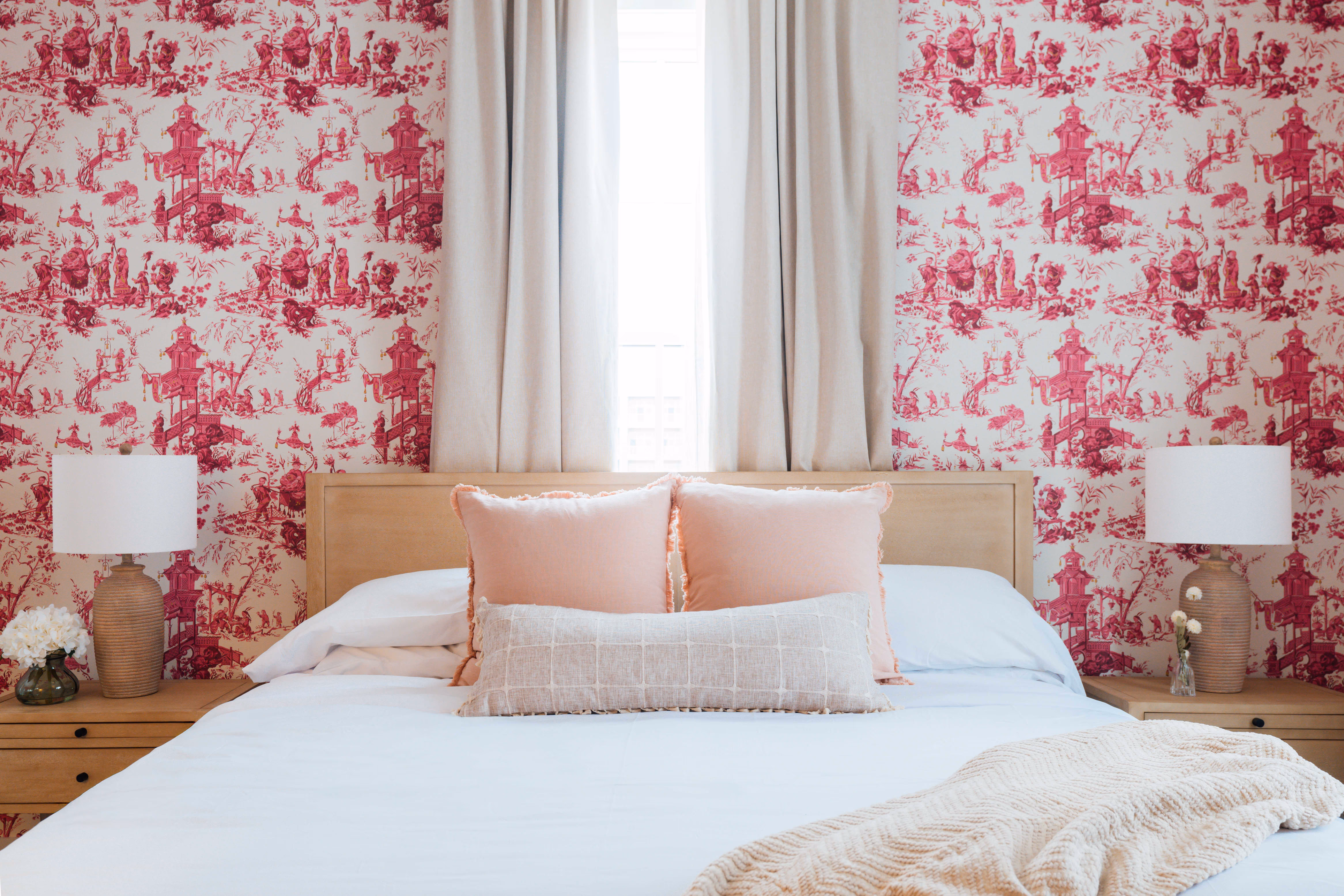 Bedroom with a wooden bed, white linens, pink and beige pillows, beige throw blanket, red patterned wallpaper, beige curtains, two wooden nightstands with matching lamps and small flower vases.