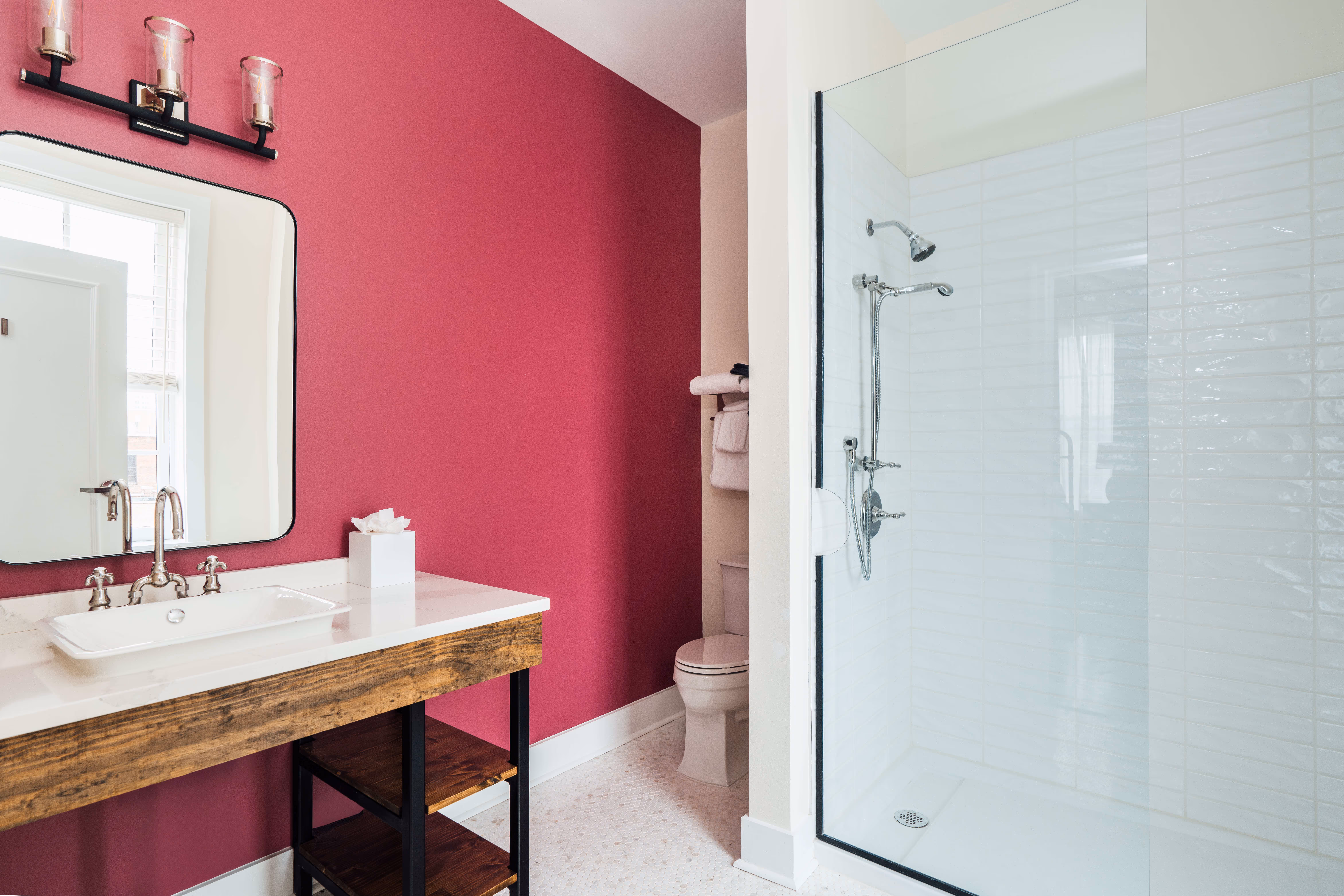 Bathroom with a white sink set in a wooden vanity against a pink wall, a large mirror, toilet with towels, and a glass-enclosed white tile shower.