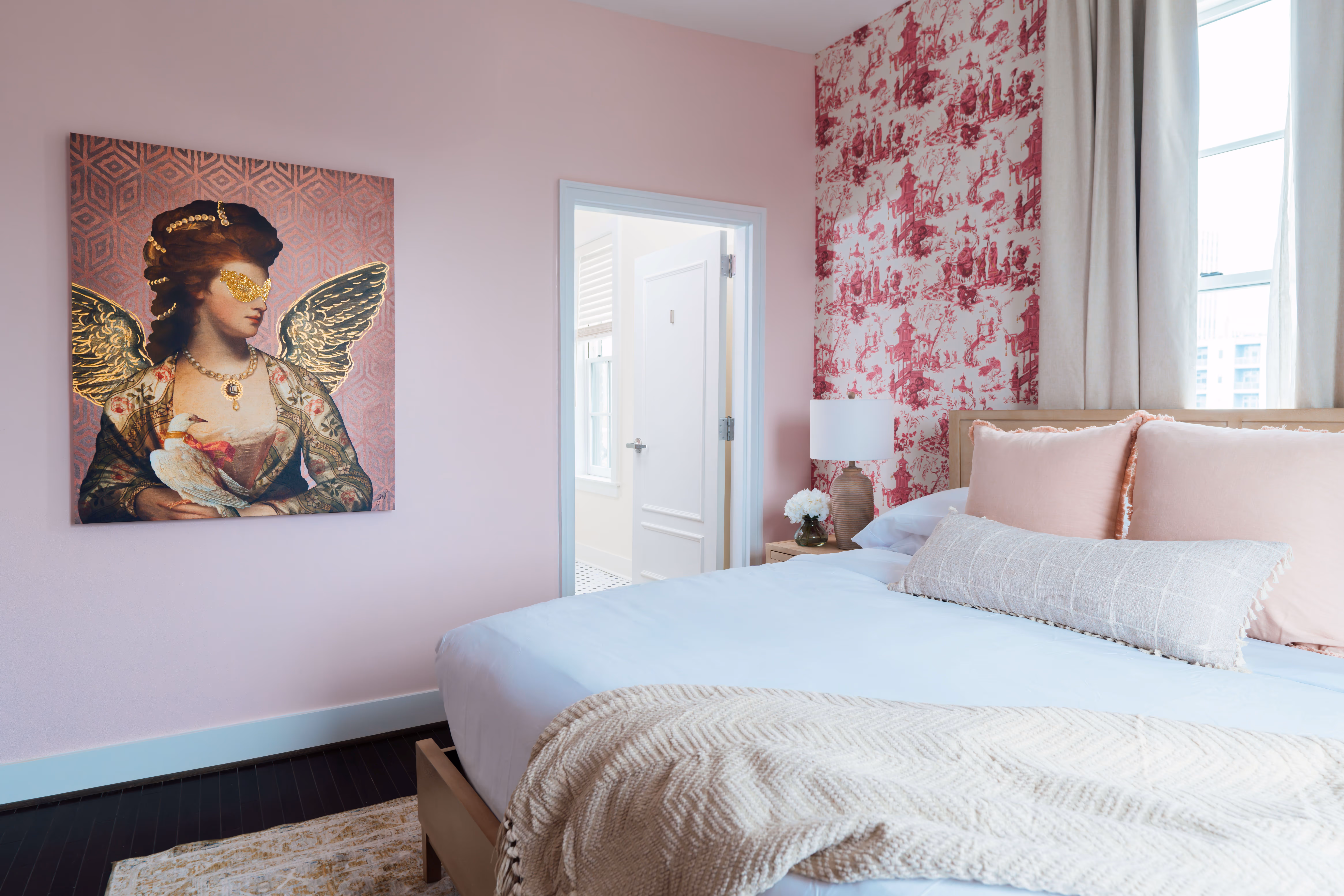 Bright bedroom with pink walls, a bed with beige and pink pillows, a patterned red wallpaper accent wall, and an artwork of a woman with golden wings holding a white bird.
