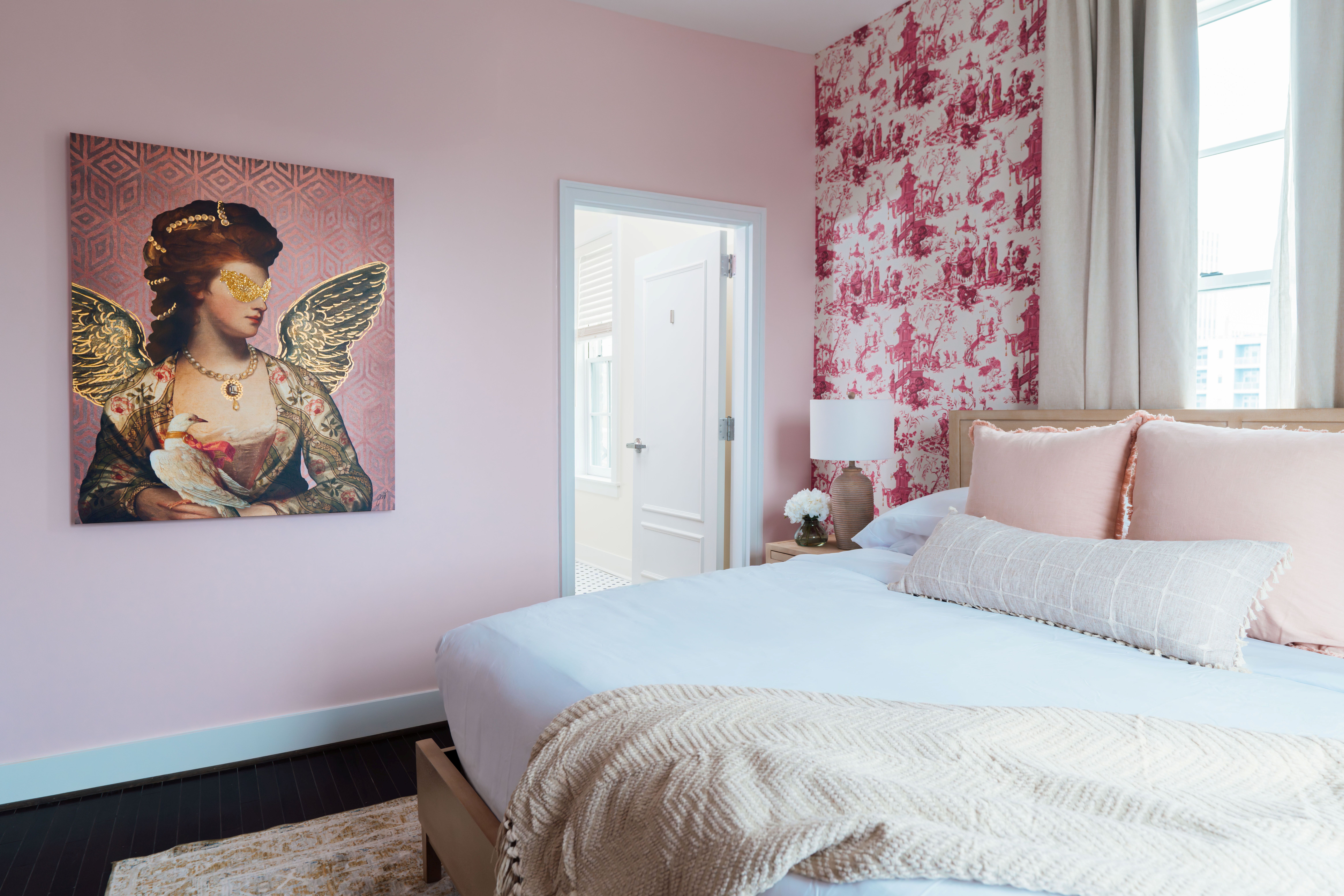 Bright bedroom with pink walls, a bed with beige and pink pillows, a patterned red wallpaper accent wall, and an artwork of a woman with golden wings holding a white bird.