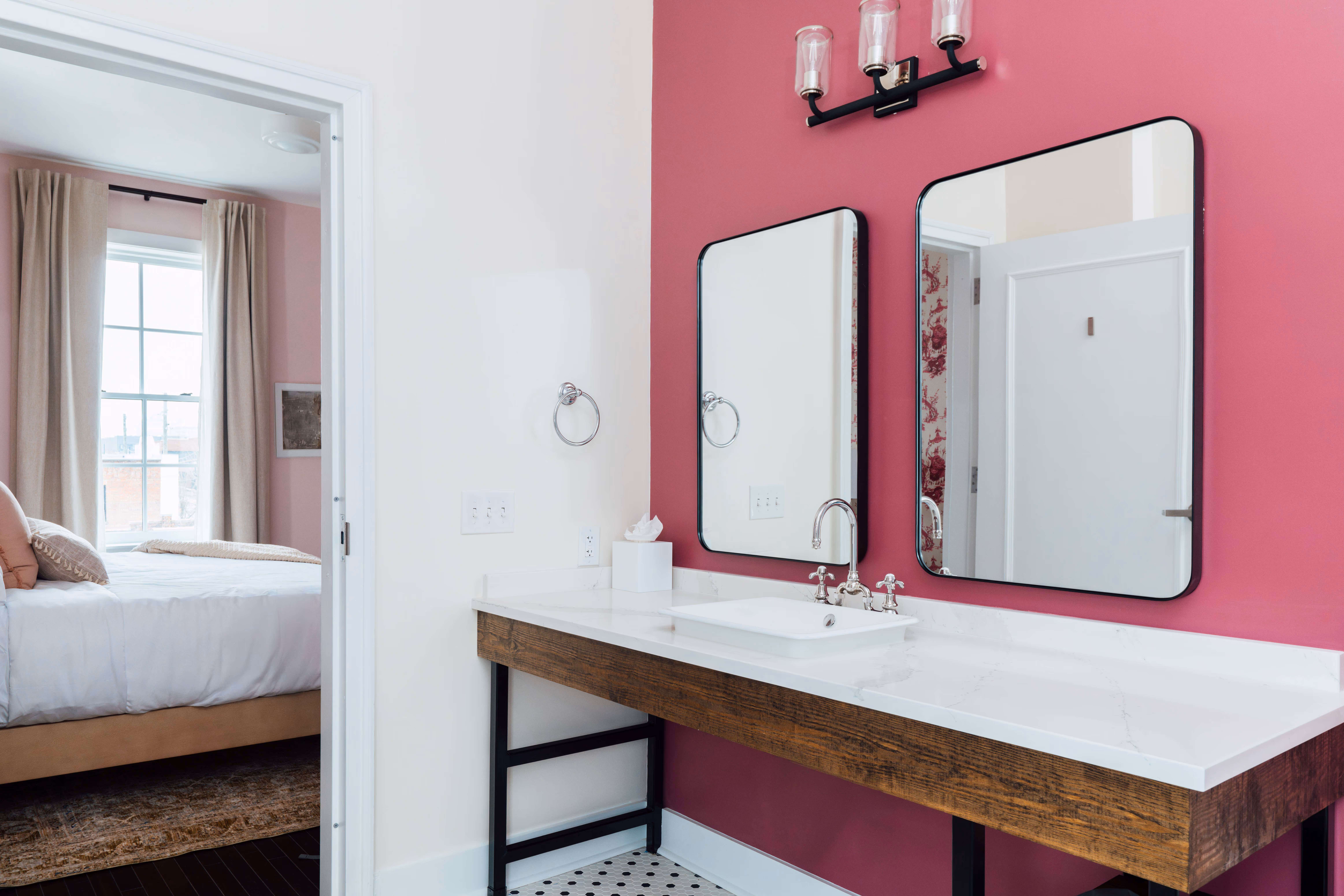Modern bathroom vanity with white sink, wooden base, two rectangular mirrors on a pink accent wall, and a glimpse into a bedroom with a bed and window.