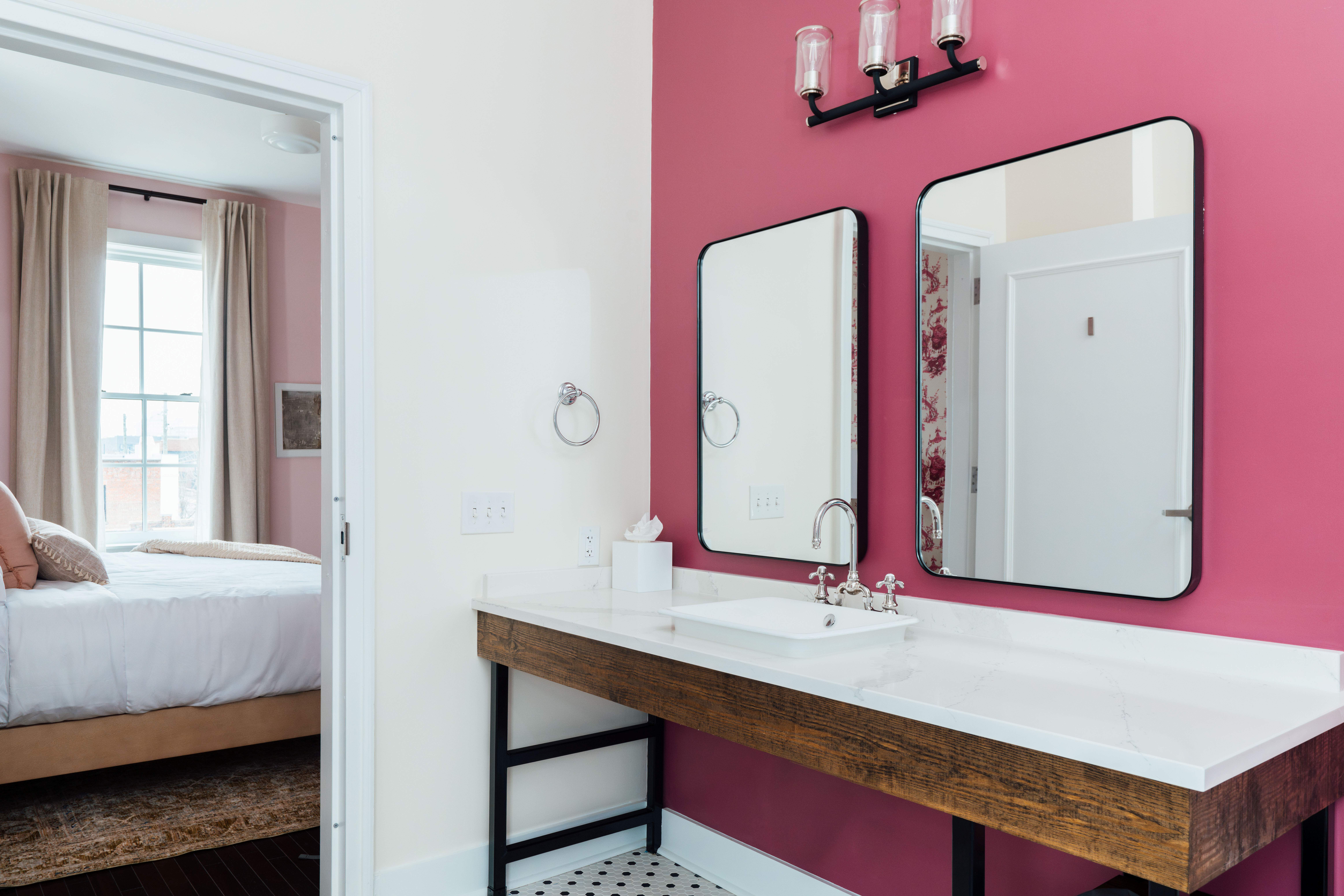 Modern bathroom vanity with white sink, wooden base, two rectangular mirrors on a pink accent wall, and a glimpse into a bedroom with a bed and window.