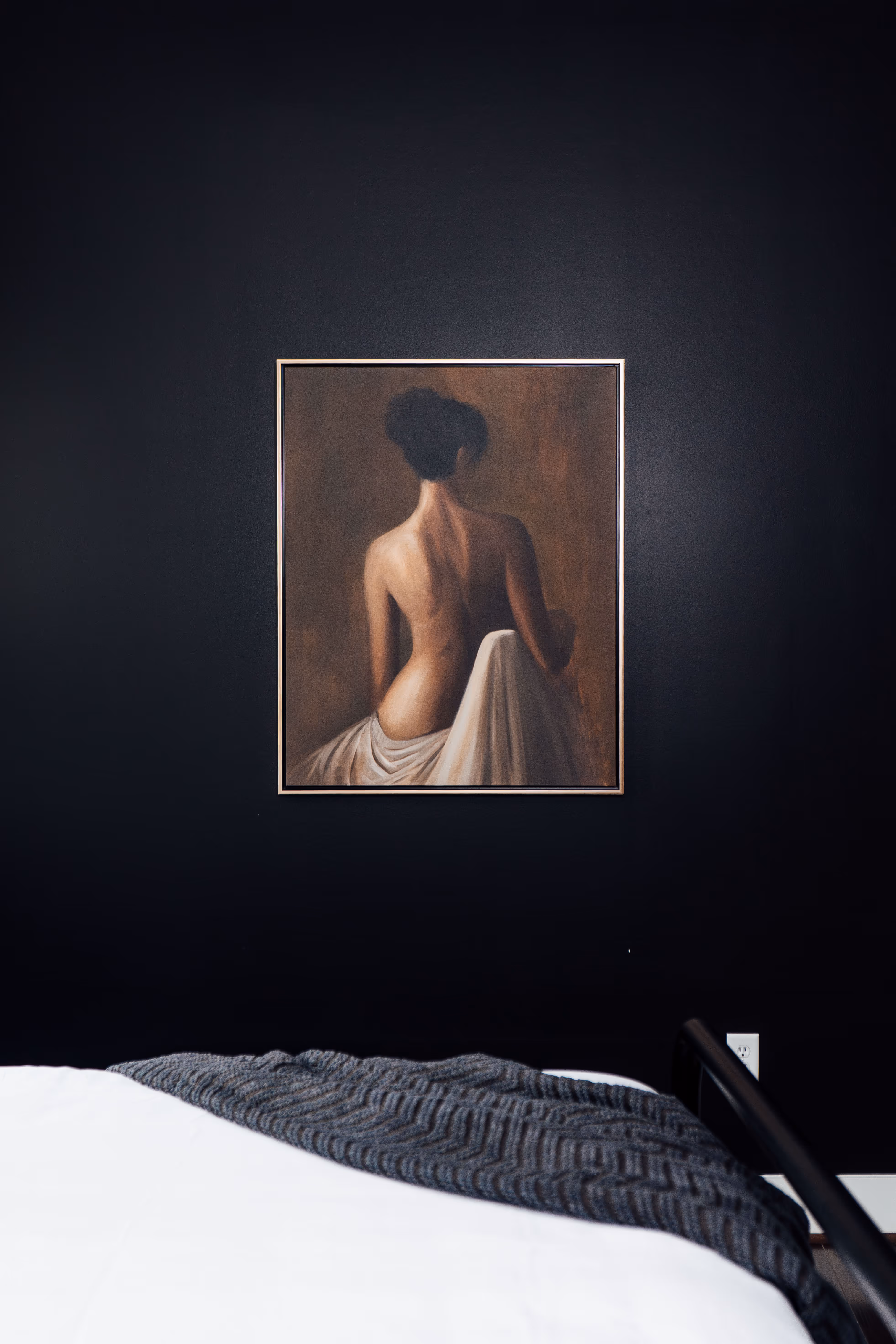 Framed painting of a woman’s bare back draped in white cloth against a dark wall above a bed with white bedding and a textured gray blanket.