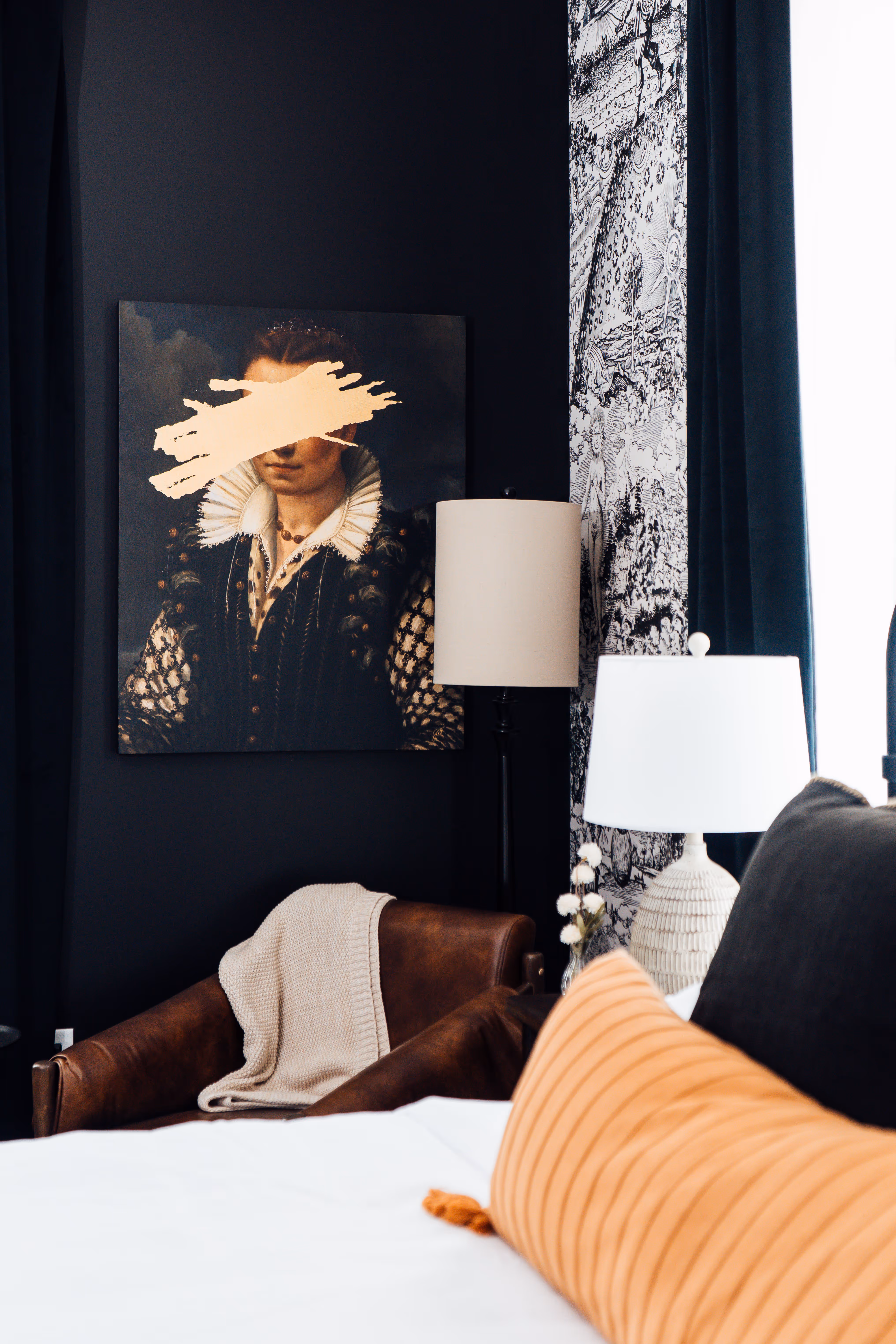 Modern room corner with leather chair draped with a cream knitted blanket, two table lamps, and a historical portrait painting with a brushstroke obscuring the eyes.