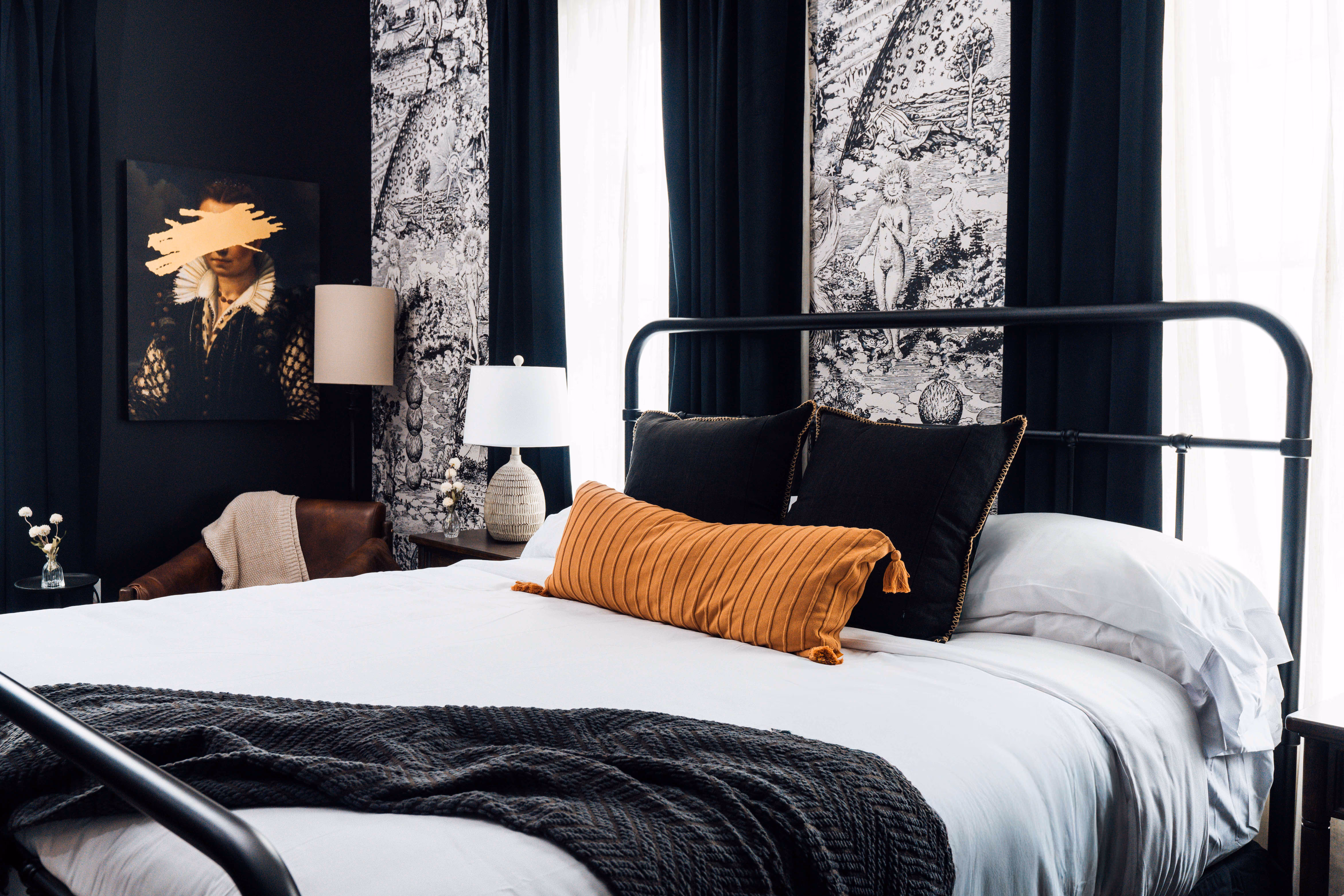Modern bedroom with black metal bed frame, white bedding, black and orange pillows, and artistic wall decor including abstract portraits and black curtains.