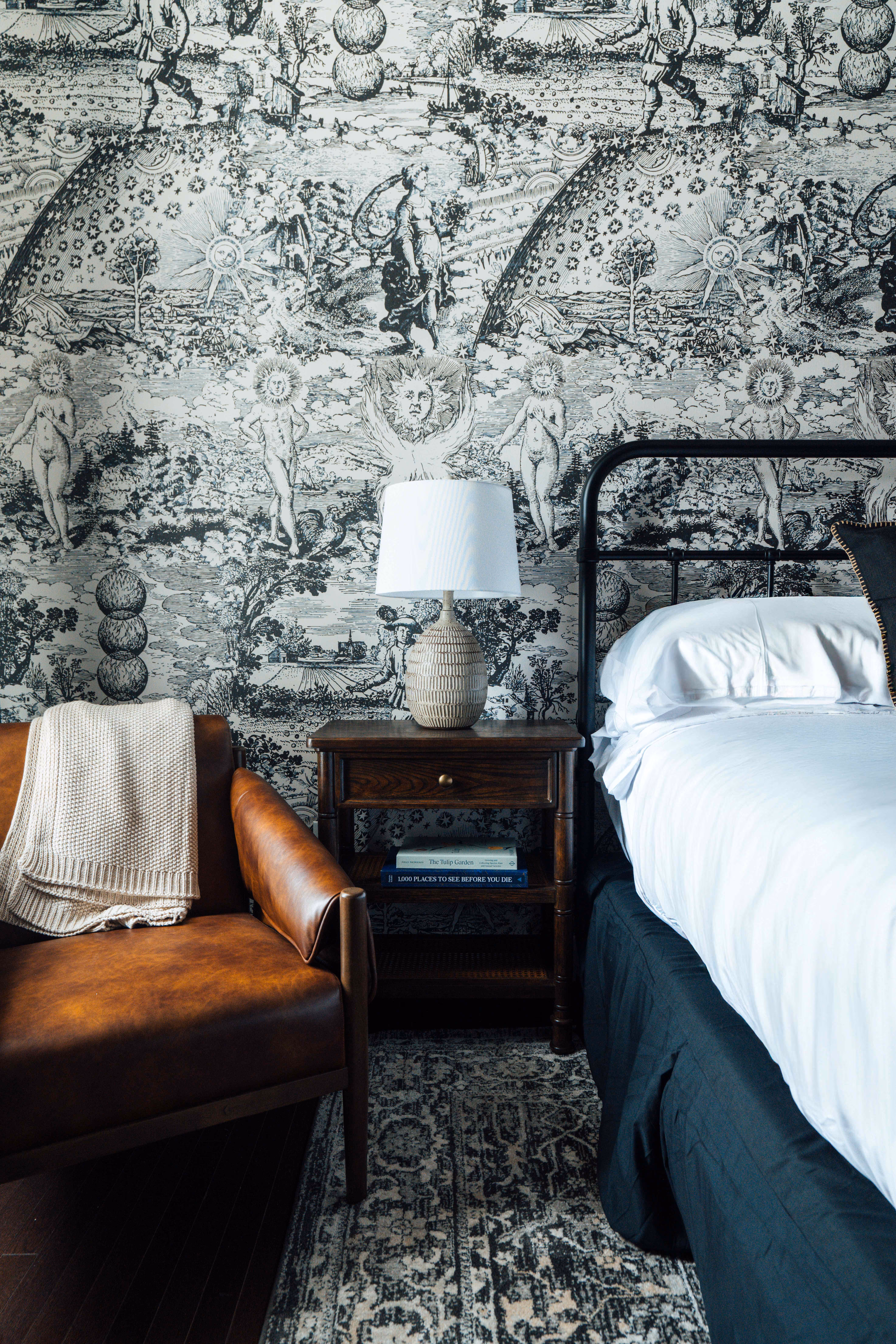Cozy bedroom corner with a brown leather armchair draped with a beige knit blanket, a wooden nightstand with a textured lamp, and a bed with white linens and a black metal frame against a black and white patterned wallpaper.