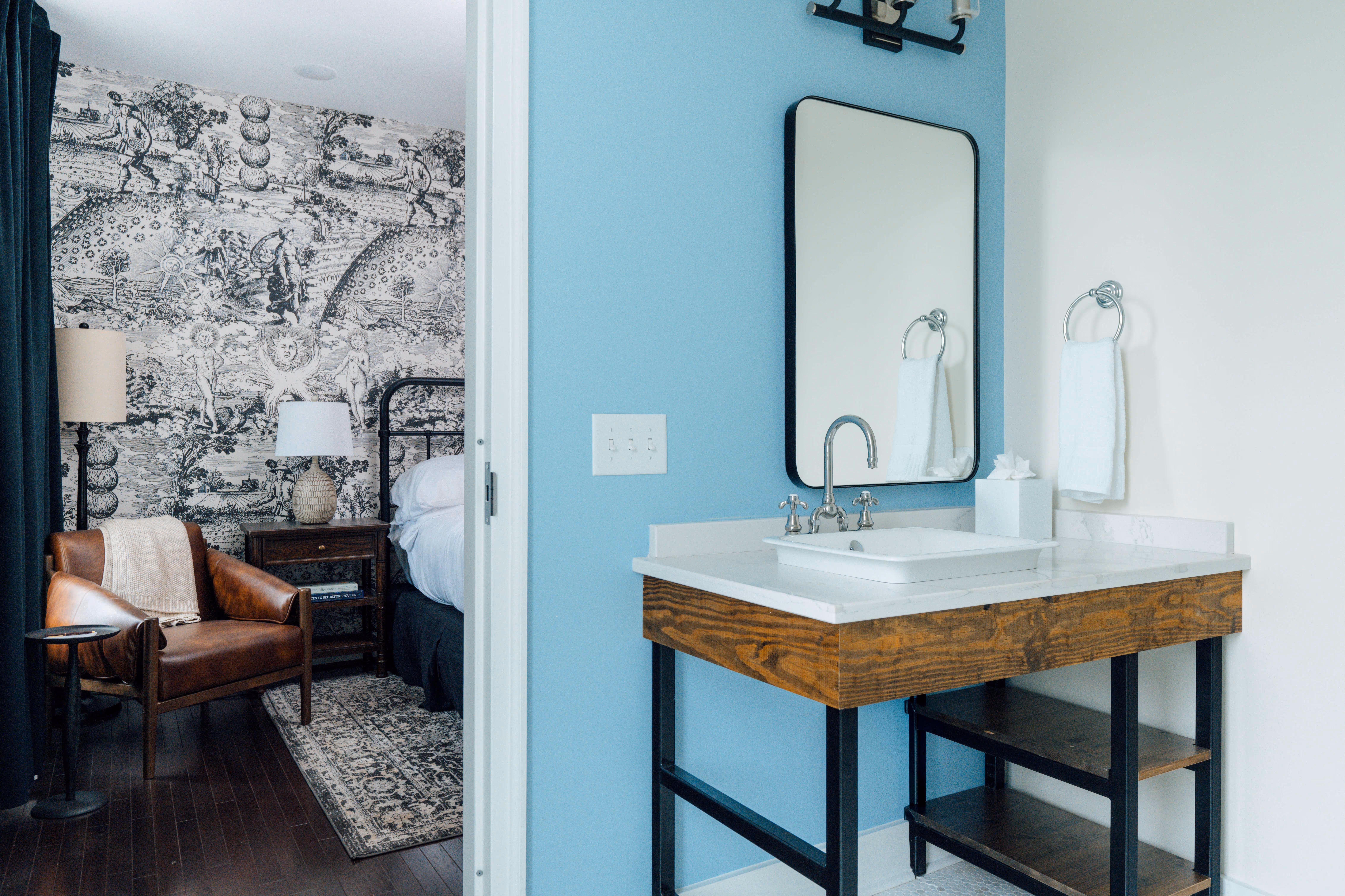 Modern bathroom sink with a rectangular mirror above and a wooden framed vanity next to a doorway leading to a bedroom with a brown leather chair and black and white illustrated wallpaper.