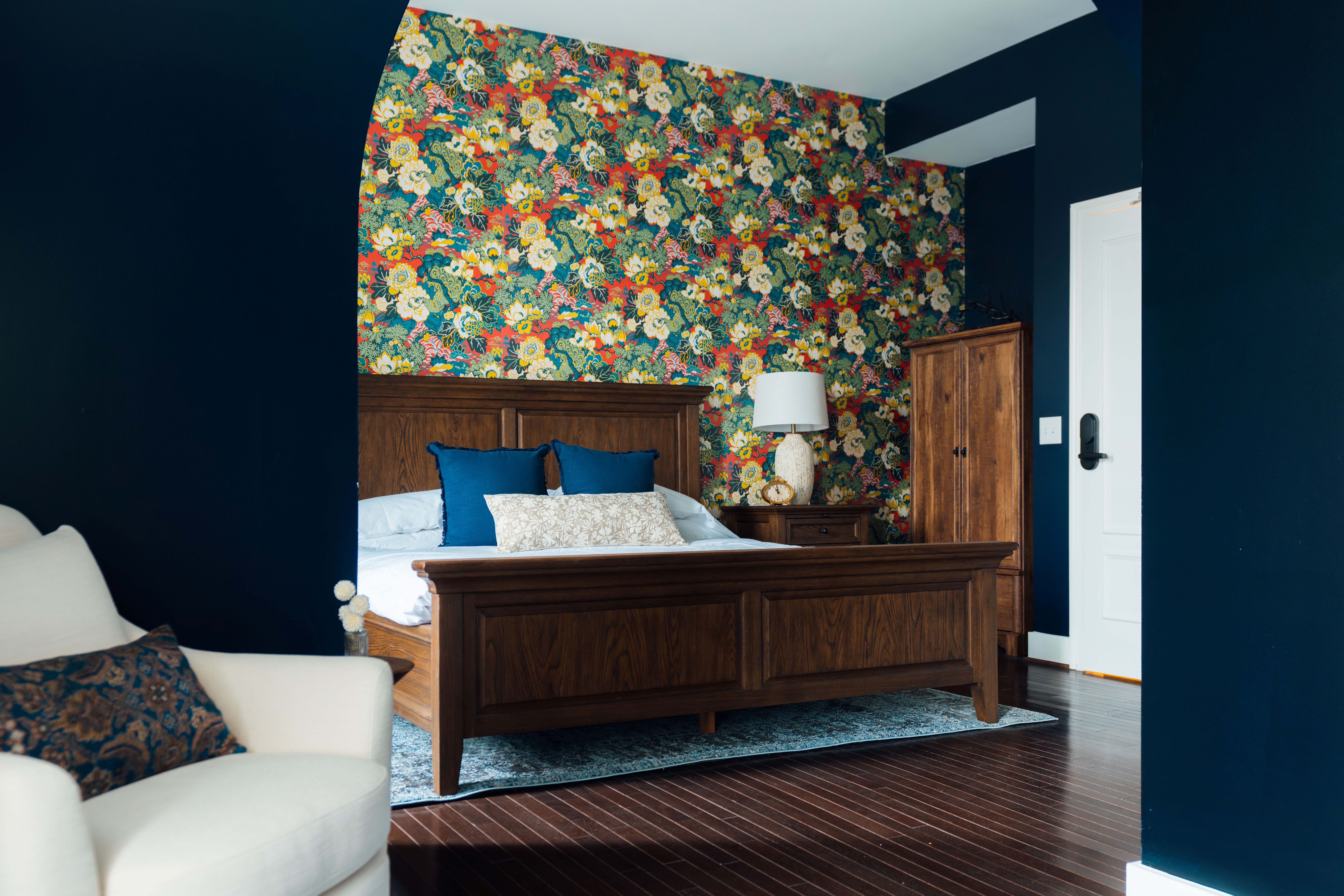 Cozy bedroom with wooden bedframe, floral wallpaper, dark blue walls, white armchair, and wooden furniture.