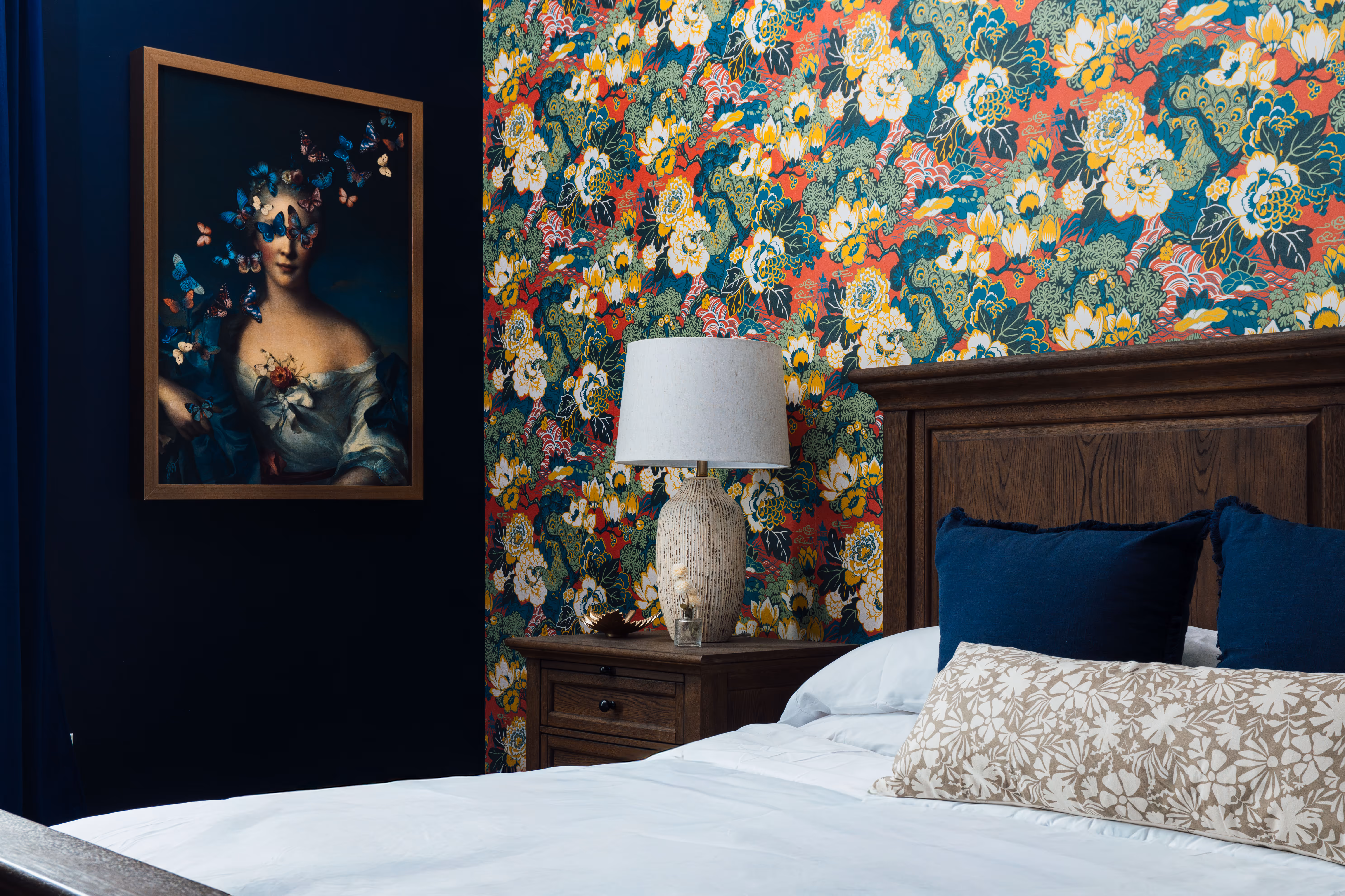 Bedroom corner with a wooden bed and nightstand, floral wallpaper, ceramic table lamp, and framed portrait of a woman with butterflies.