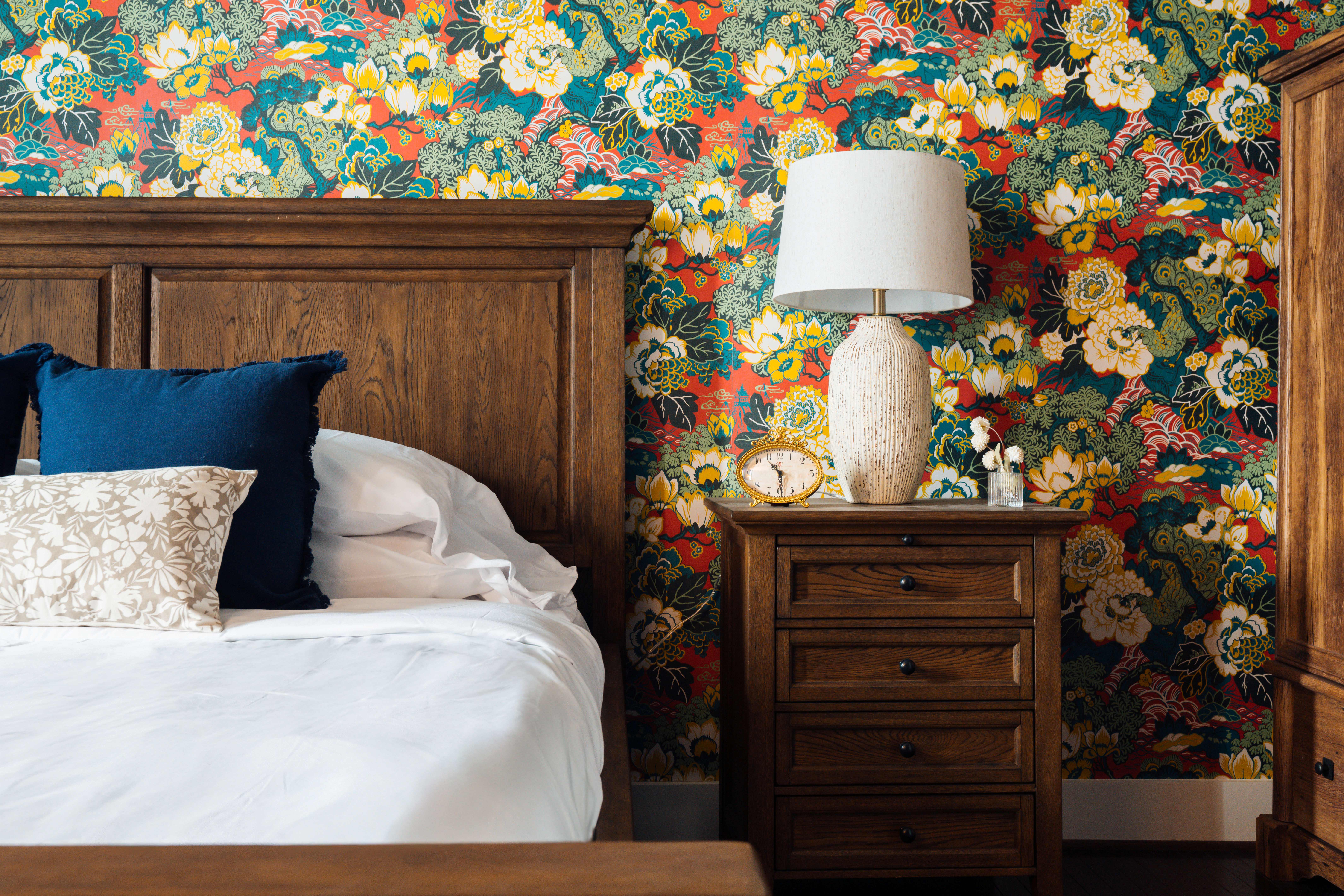 Bed with wooden headboard, white bedding, blue and beige pillows, next to a wooden nightstand with a white lamp, vintage clock, and small vase against floral wallpaper.