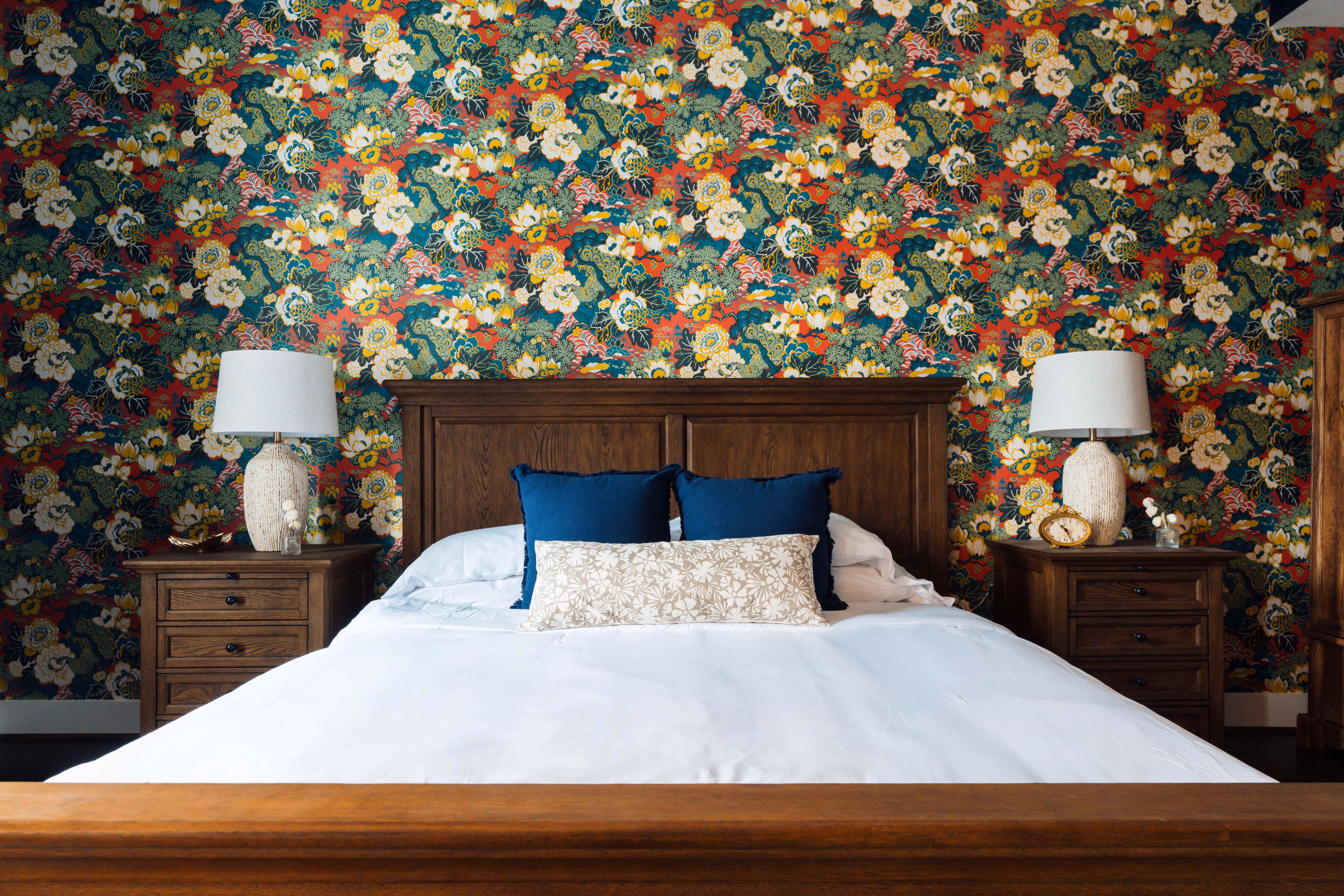 Wooden bed with white bedding and blue accent pillows against a colorful floral wallpaper, flanked by matching wooden nightstands with lamps.