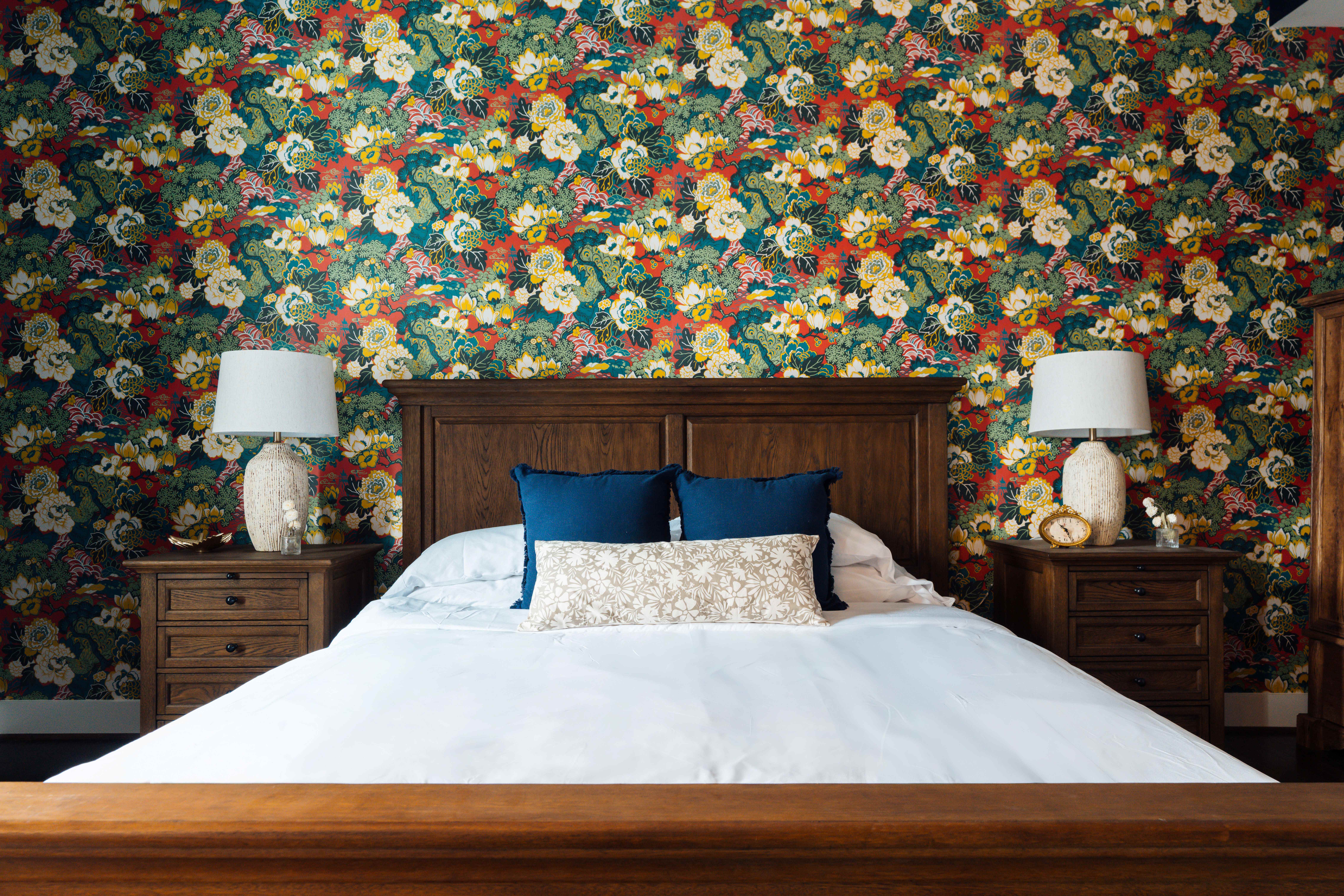 Wooden bed with white bedding and blue accent pillows against a colorful floral wallpaper, flanked by matching wooden nightstands with lamps.