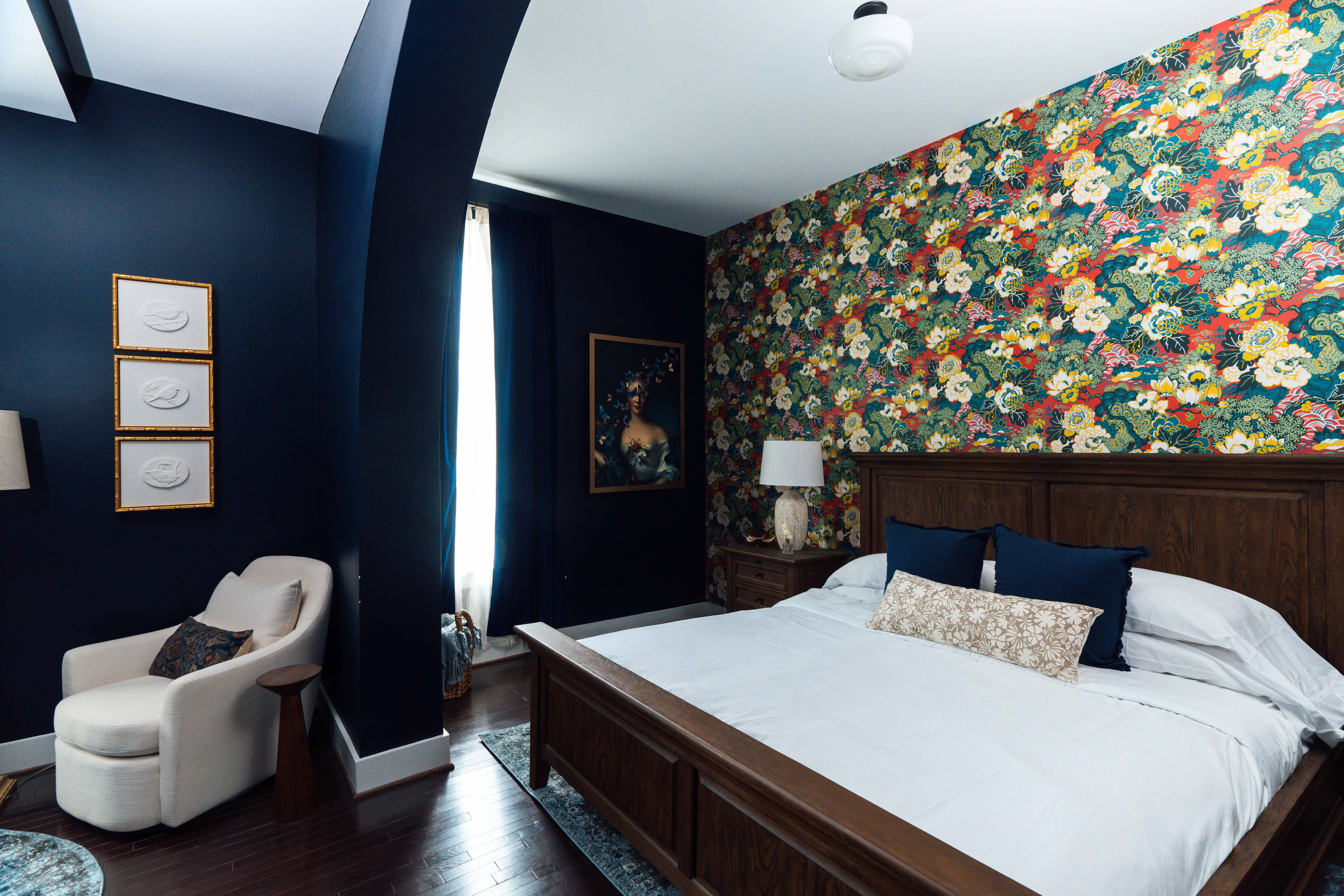 Bedroom with dark blue walls, a floral patterned accent wall, wooden bed with white bedding and blue pillows, a white armchair, and framed artwork.